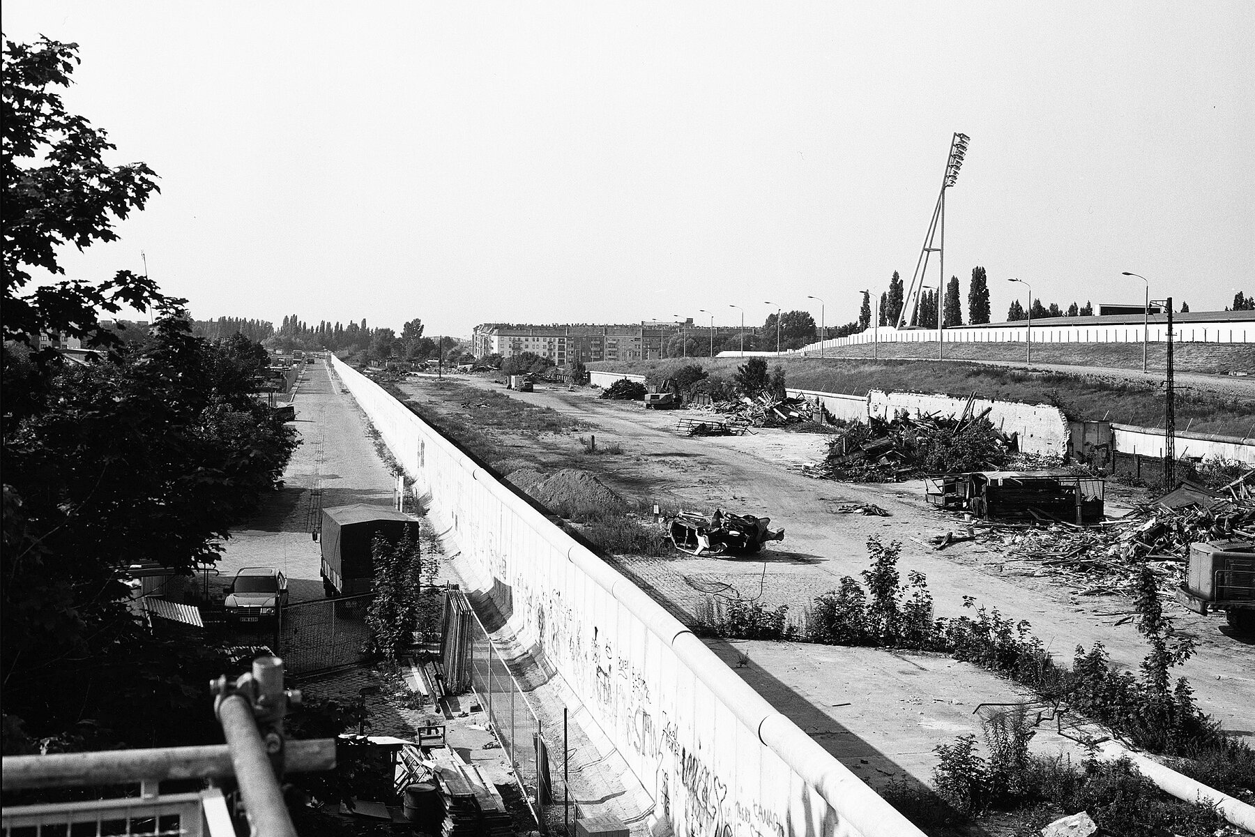 Die Berliner Mauer im späteren Gelände des Mauerparks. Rechts der Mauer befindet sich Schutt, ganz rechts sind die Flutlichter des Jahnsportparks. 