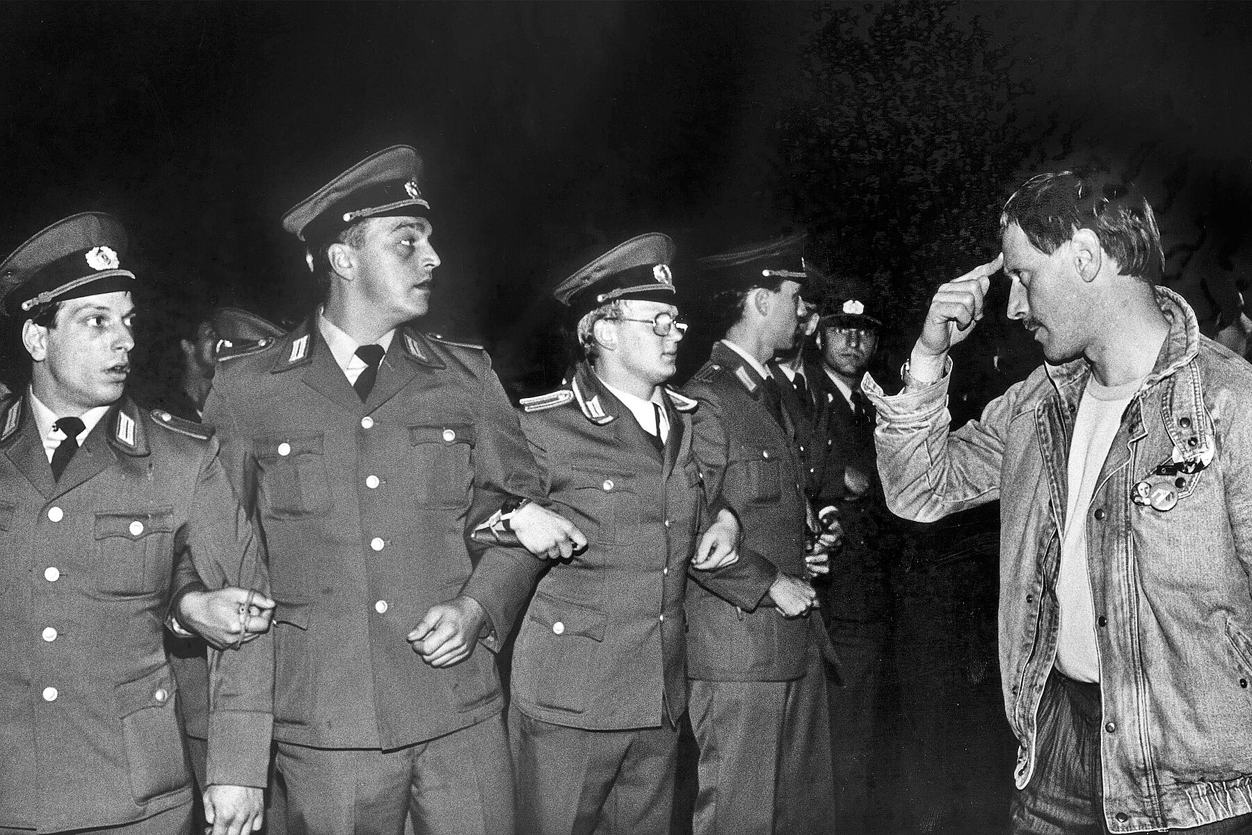 Four men in uniforms stand in a row. Their arms are interlocked. A man gestures that they are crazy by pointing his index finger to his head. rm stehen in einer Reihe. Ihre Arme sind untereinander verschränkt. Ein Mann rechts zeigt ihnen einen Vogel. 