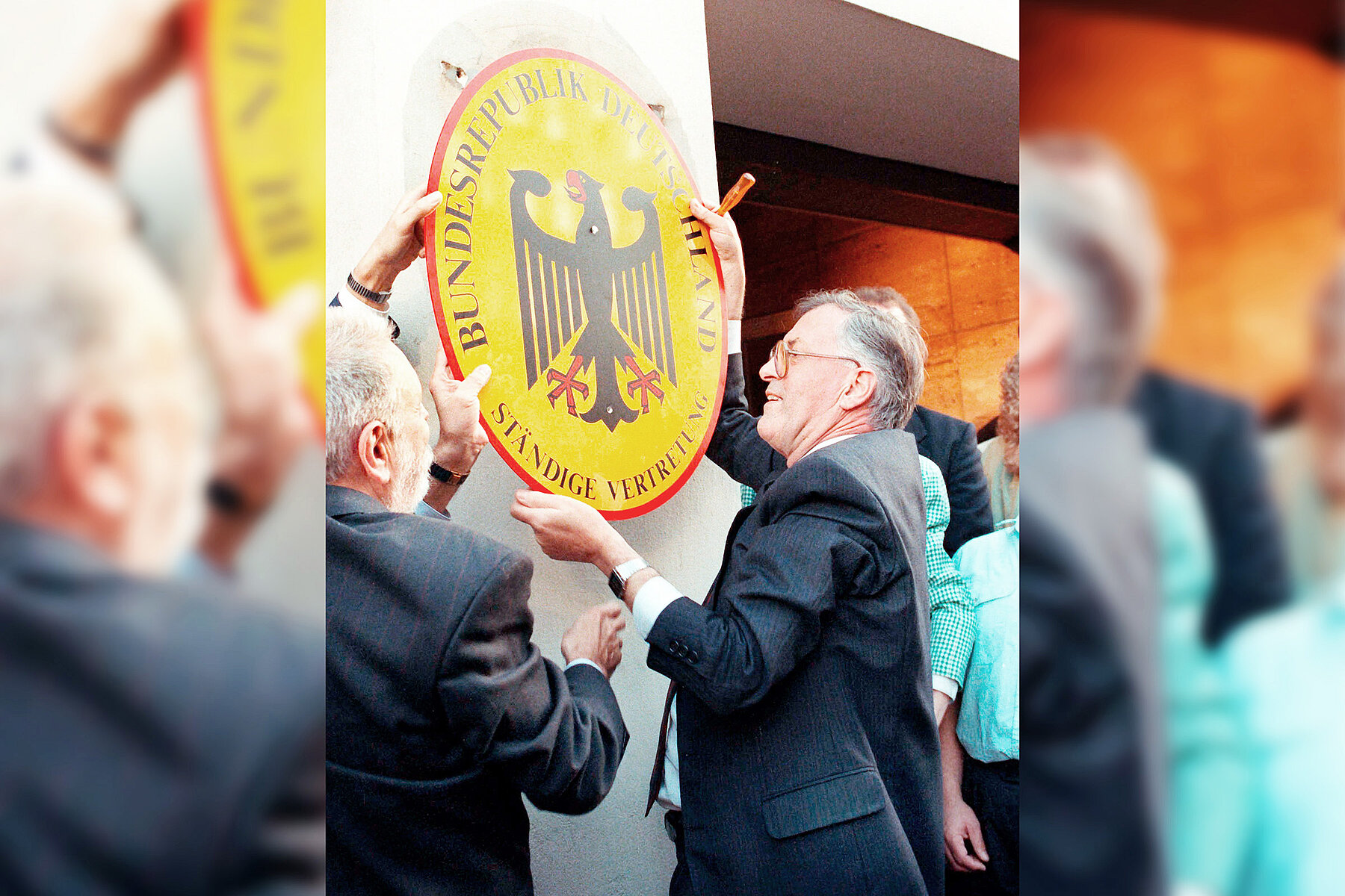 Der Leiter der Ständigen Vertretung hängt mit einem weiteren Mann das Amtsschild mit schwarz-rotem Bundesadler auf gelben Grund ab. 