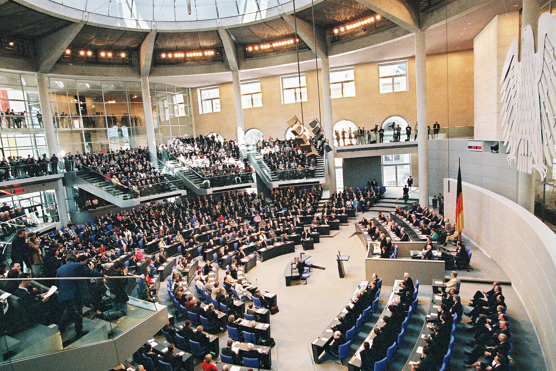 Ansicht von oben auf den Plenarsaal mit voll besetzten Stuhlreihen, die kreisförmig angeordnet sind. Rechts hängt ein Bundesadler an der Wand. 