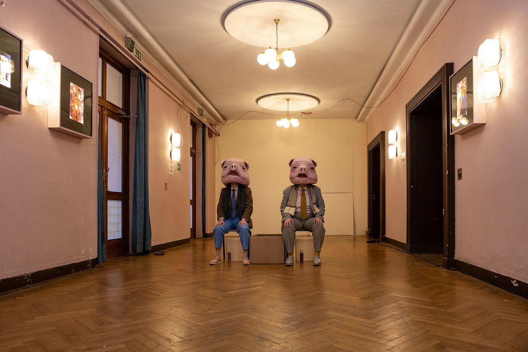 Two people with pig masks on their heads sit side-by-side on chairs in the middle of a room with a parquet floor.