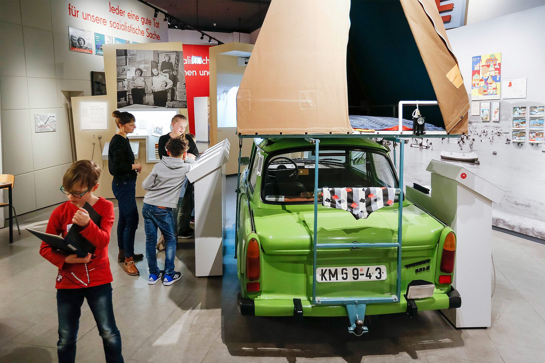 The exhibition of the museum in the Kulturbrauerei. On the left side of the picture are children, on the right side is a green Trabi with a tent on the roof.