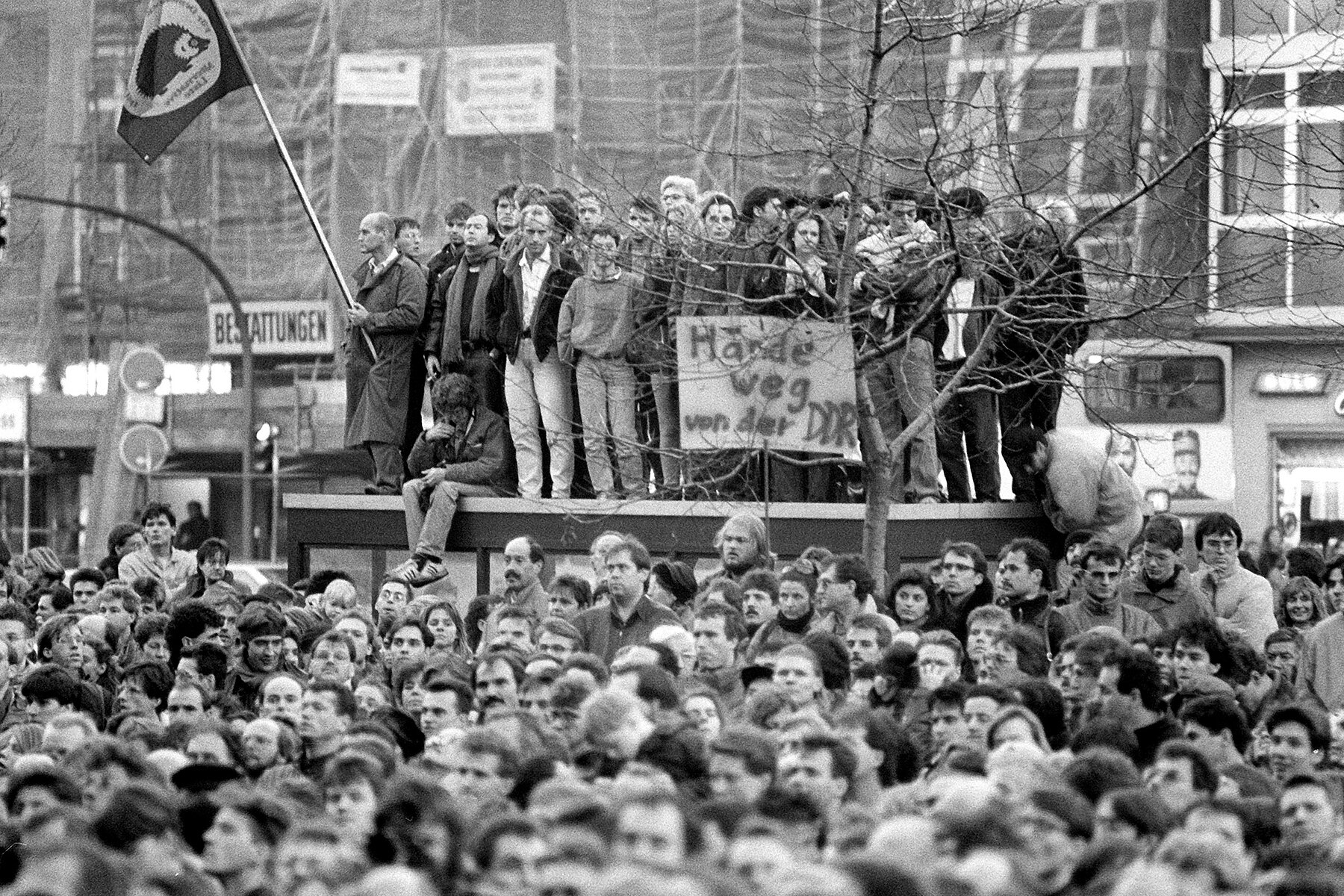 Demonstrators in front of a house with a construction fence. Some stand on top of a building and hold a sign which reads: Hands off the GDR. 