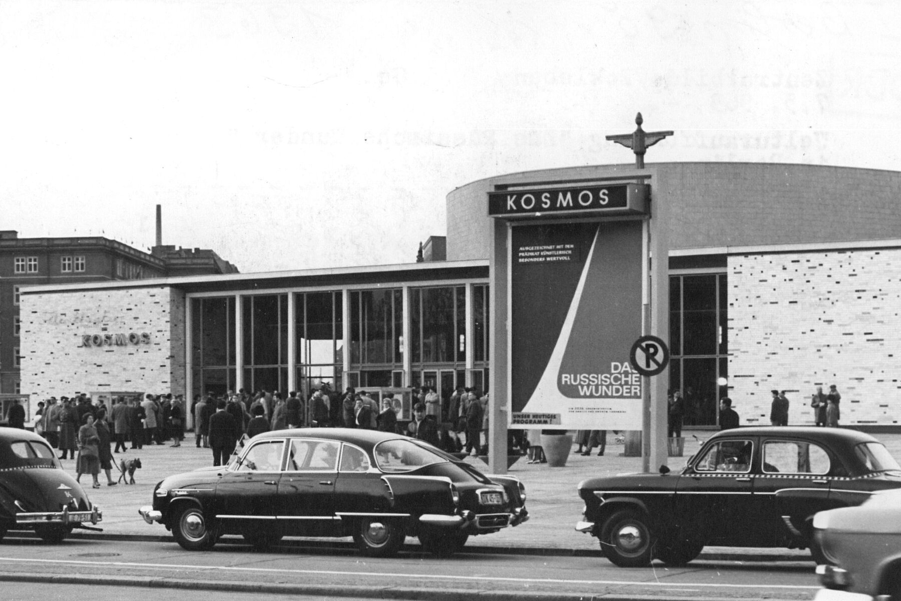 Exterior shot of the Kosmos Cinema building with a rotunda in the middle. There are many people and three cars in front of the cinema.