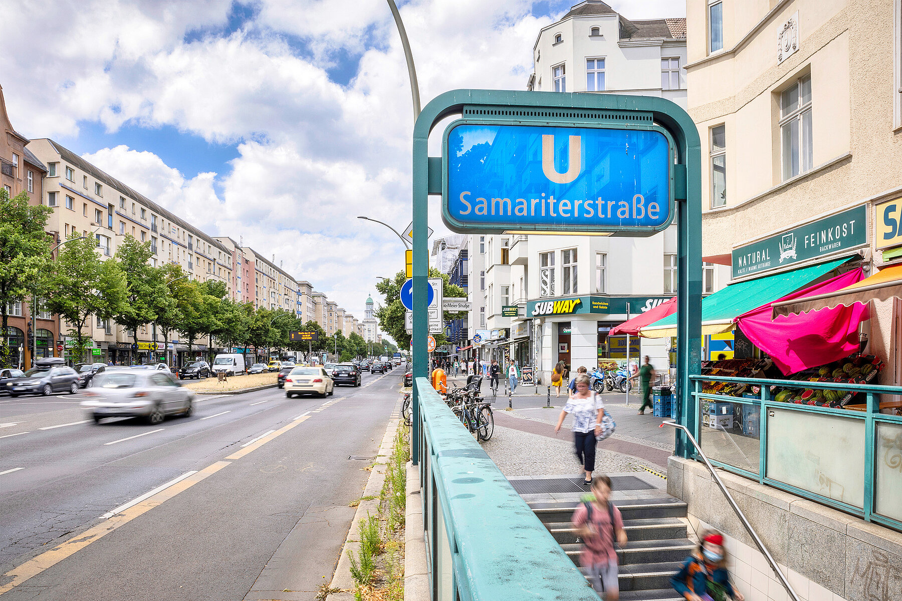 Rechts überspannt ein blaues Schild mit der Aufschrift U Samariterstraße eine Treppe, die in die Haltestelle hinab führt, links davon eine Straße. 