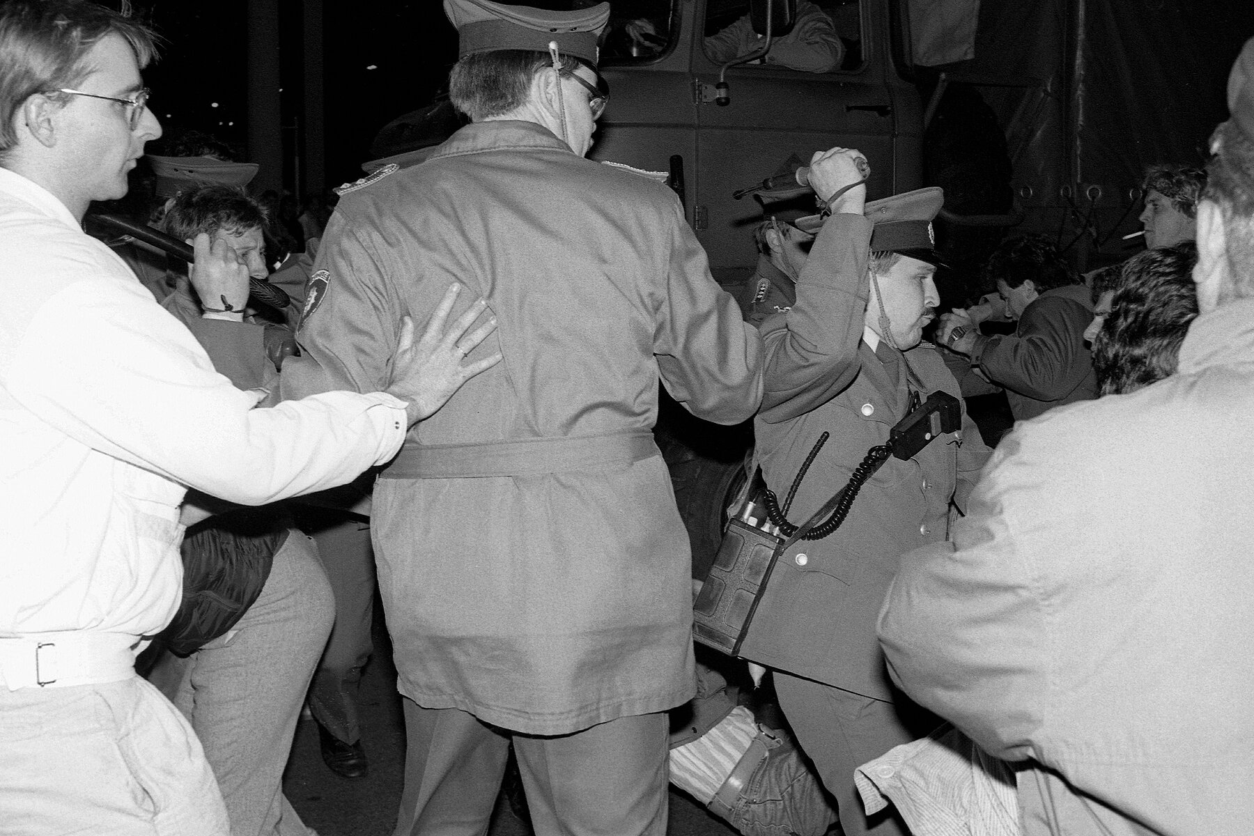 Several uniformed police officers hold up batons. On the left, a man touches a policeman on the back with his hand.