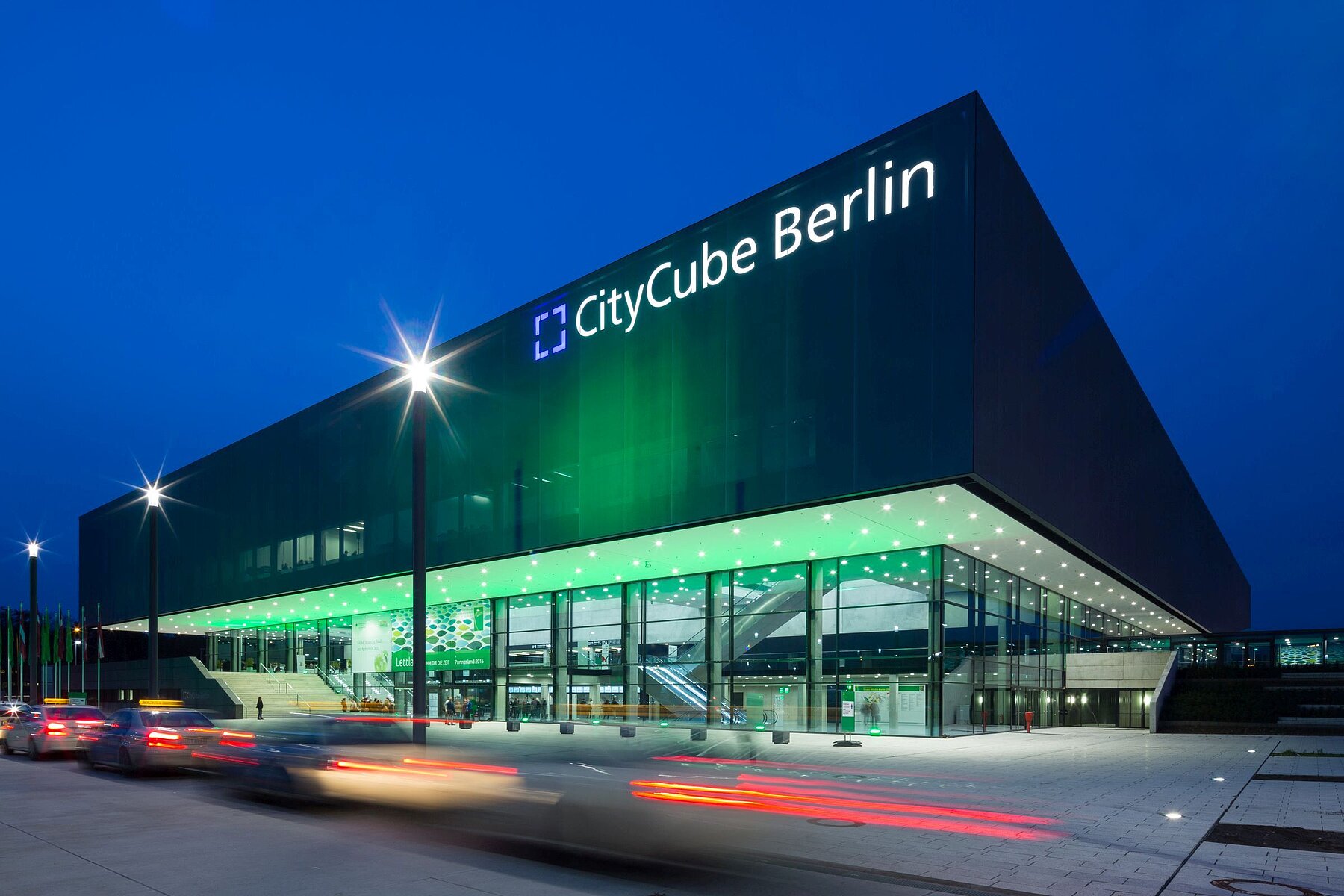 Der City Cube Berlin, nachts grün beleuchtet. Vor dem Gebäude parken Taxis.
