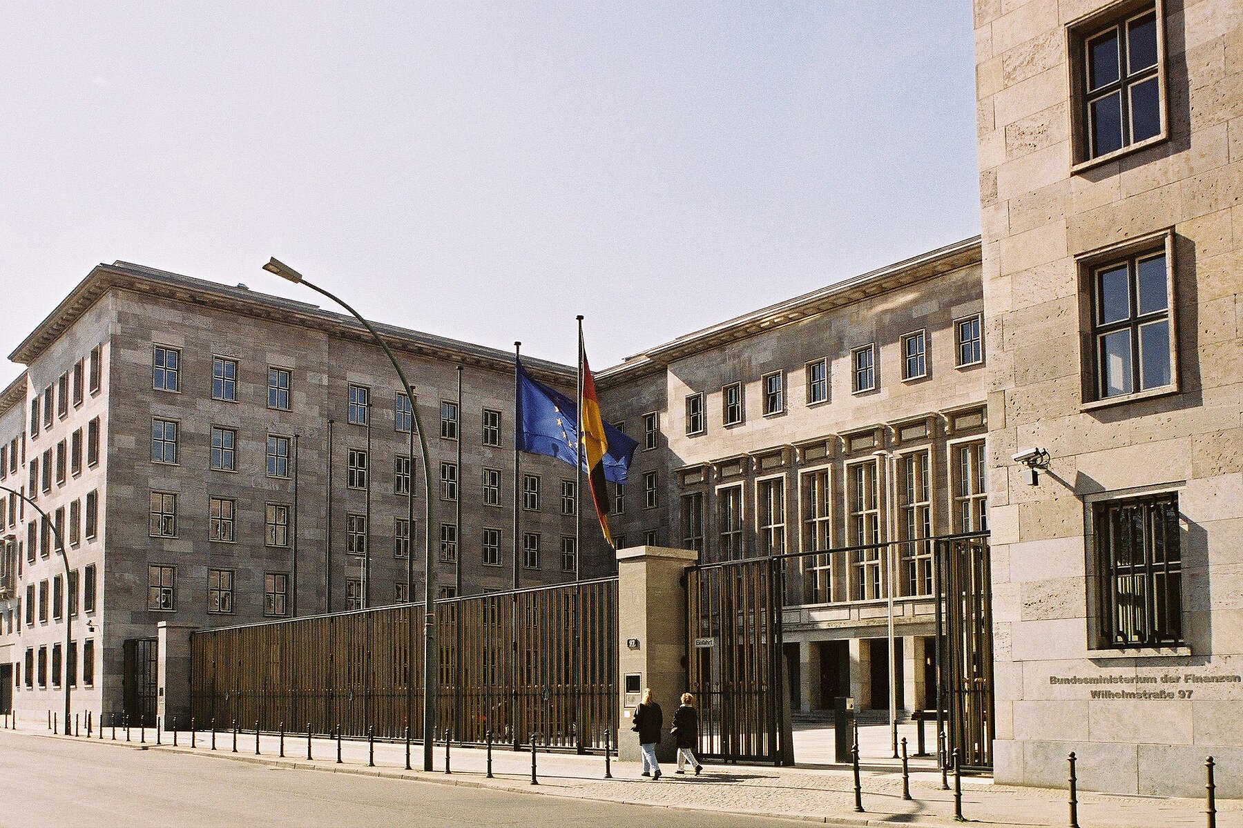 View of a grid fence in front of the Federal Ministry of Finance. In front of the builiding a European and a German flag are waving in the wind.