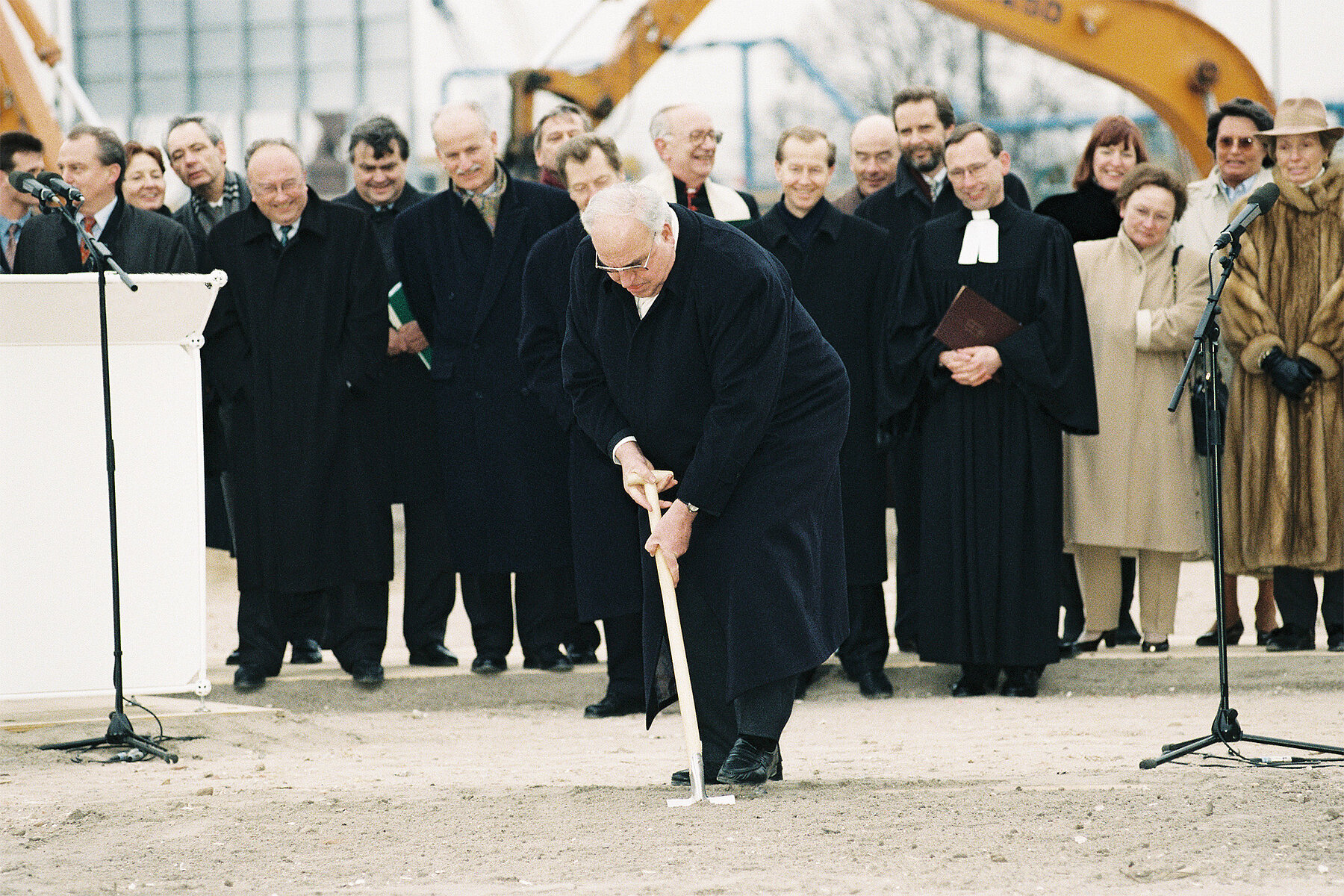 Helmut Kohl stabs the ground with a spade, behind him stands a row of people, including a pastor holding a Bible.