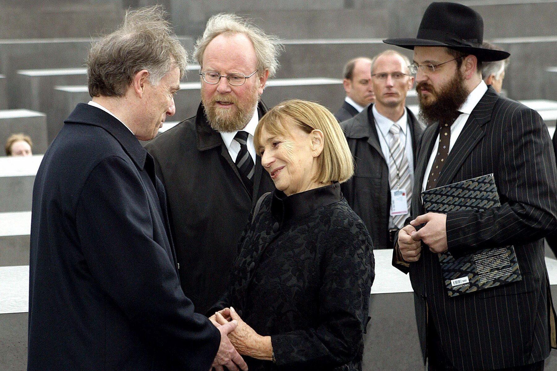Two politicians, a woman and a Rabbi, all in black clothing, in a conversation in front of the concrete stelae of the memorial.