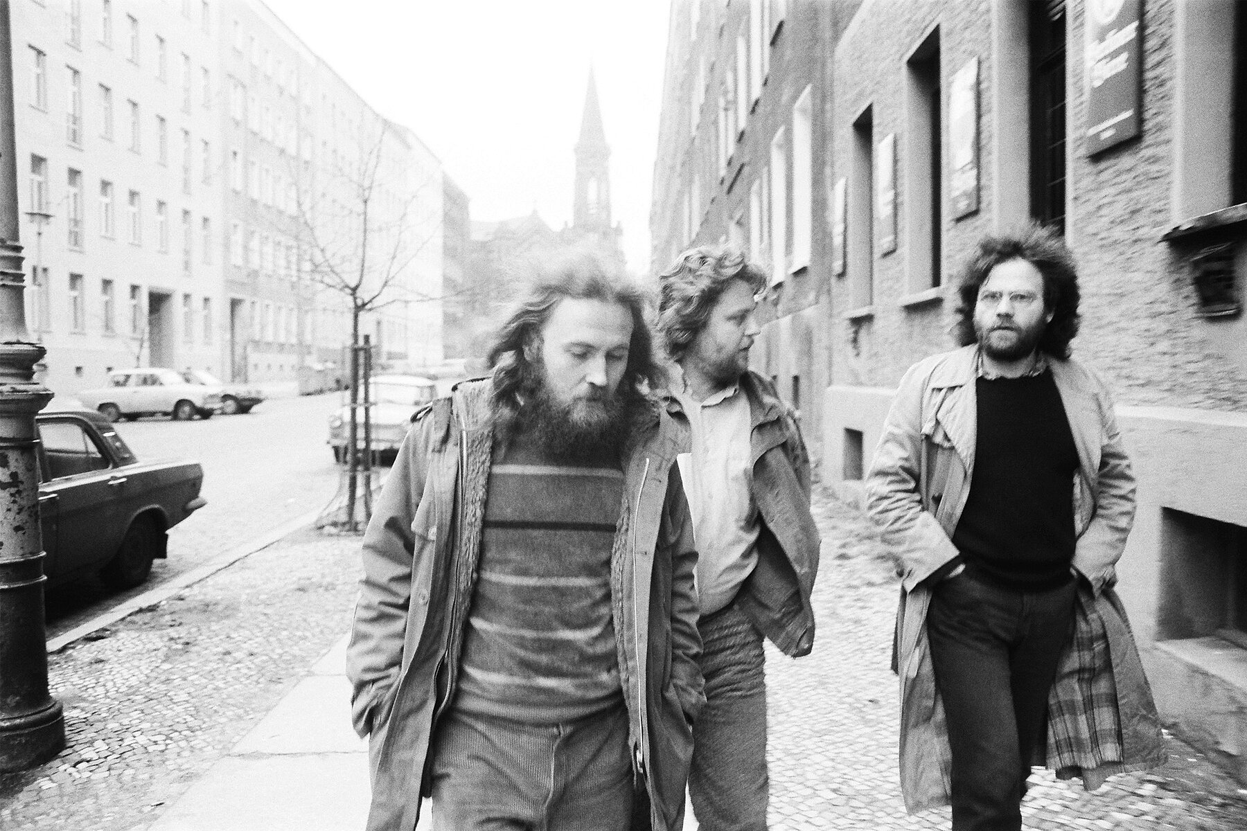 Three men walk down a street, on the left and right are apartment buildings. In the background is a church steeple. 