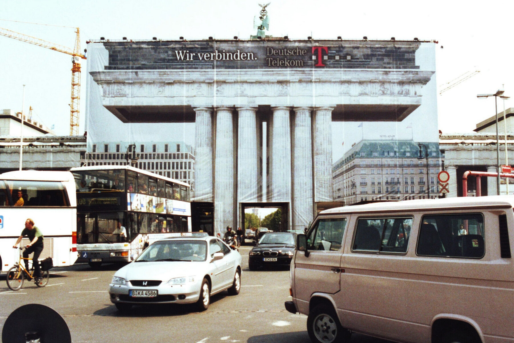 Das Brandenburger Tor ist mit einem Werbebanner der Deutschen Telekom verhüllt. Darauf sind die angrenzenden Gebäude und Teile des Tores abgebildet. Durch die Mitte fahren Autos.