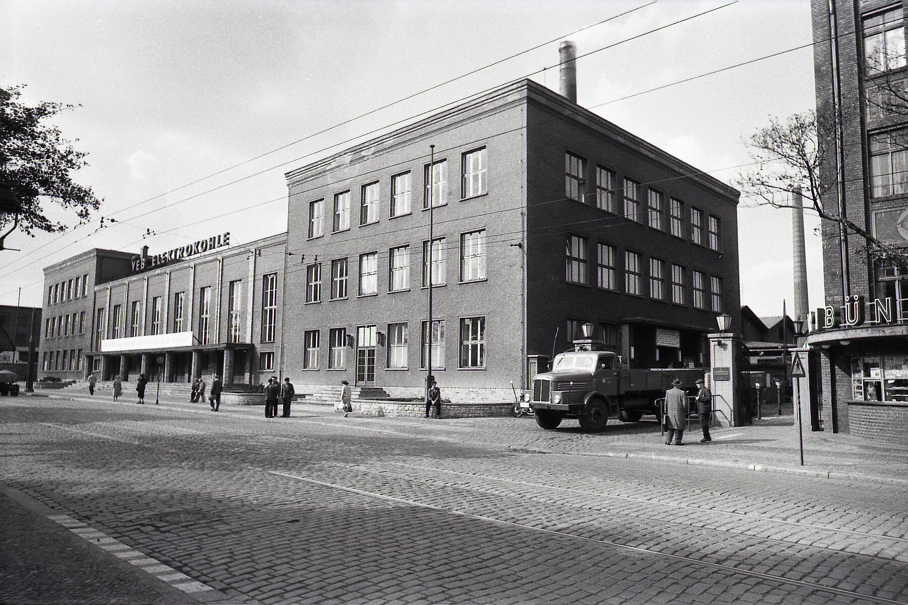 Kopfsteingepflasterte Straße mit einem großen Backsteinindustriegebäude im Hintergrund. Davor laufen Personen. Rechts des Gebäudes fährt ein Auto aus einer Einfahrt. 