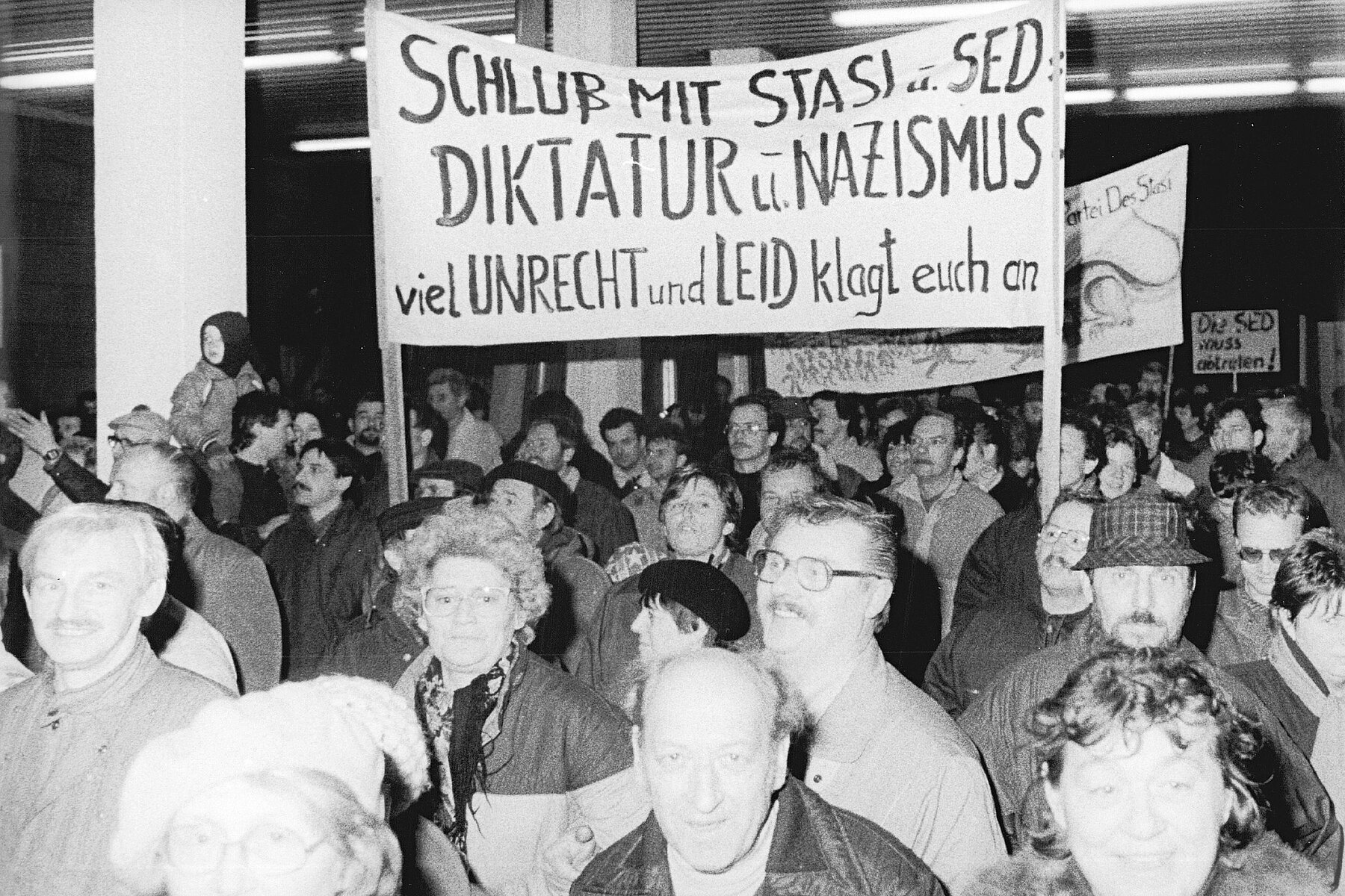 Several people with protest banners in the building. One sign reads: End Stasi and SED, dictatorship and Nazism, much injustice and suffering accuse you. 