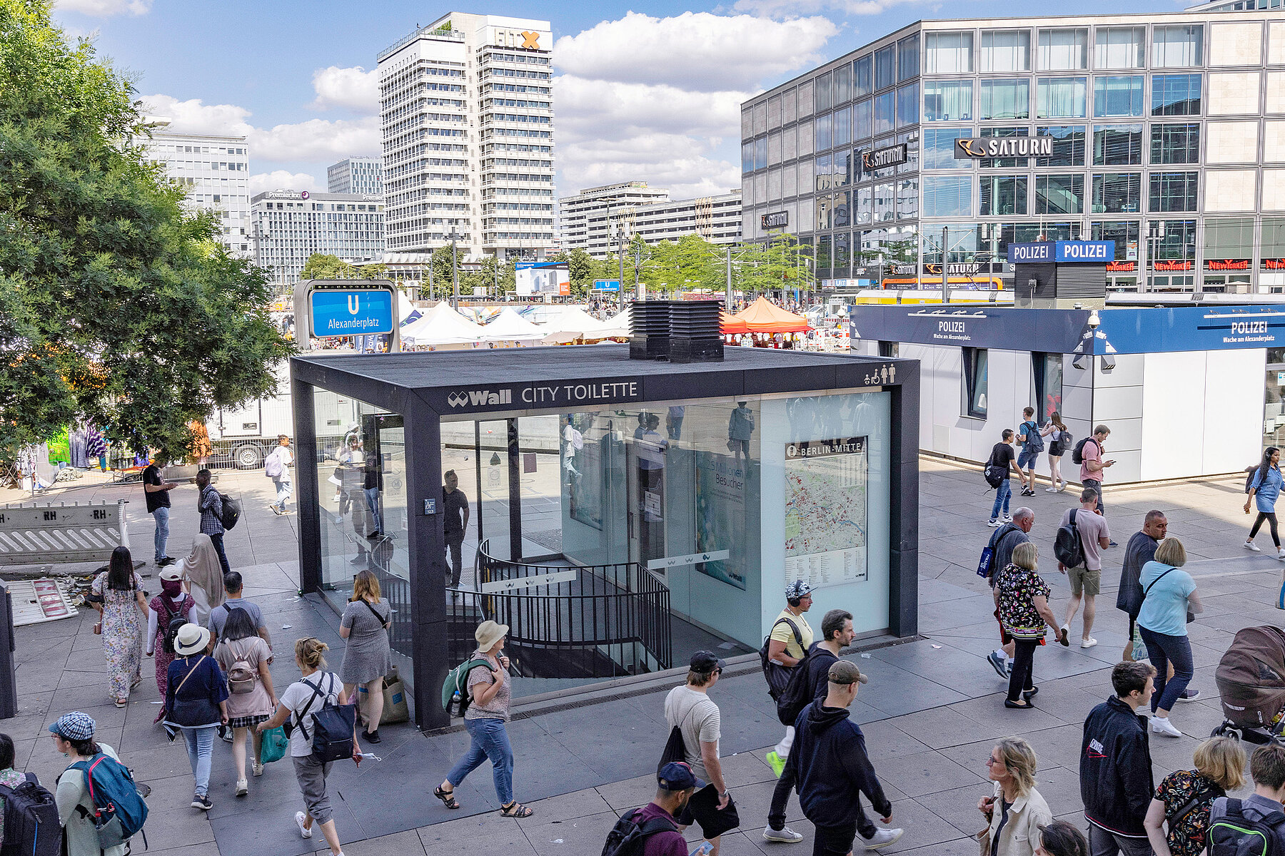 People walk across Alexanderplatz. On the left a public toilet building, on the right a small police station.