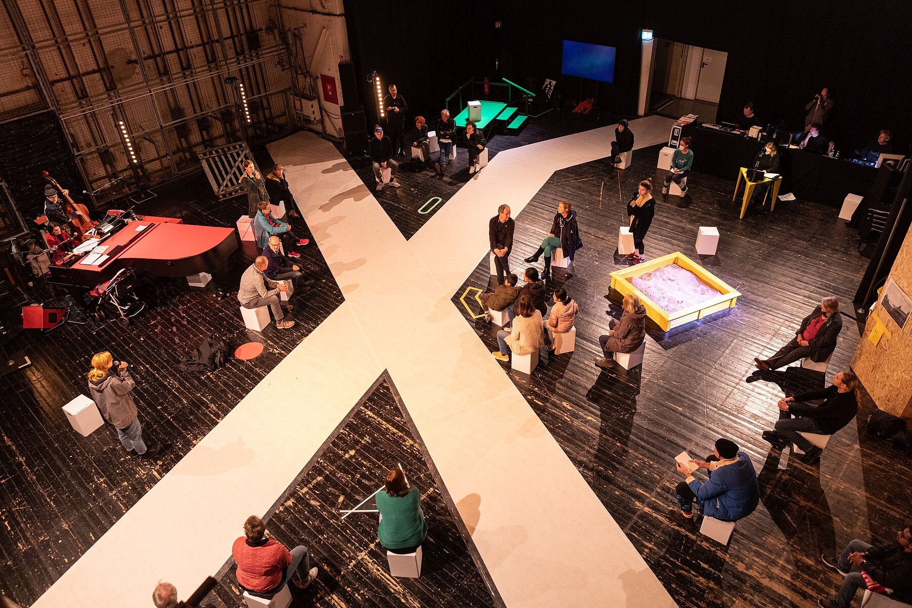 A white X the size of a room lies on the floor, with people sitting on chairs around it.