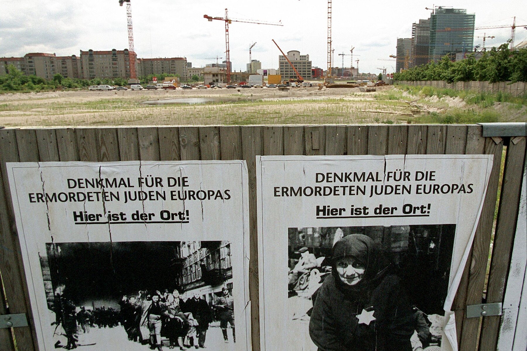 In the foreground is a construction fence made of wood with two posters. Next to historical pictures of Jewish persons it reads: Memorial to the Murdered Jews of Europe. This is the site. In the background is a meadow, behind it are buildings and construction cranes. 