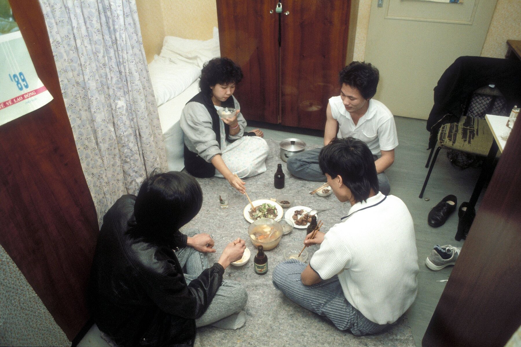 Four people sit in a circle on a carpeted floor in a small room and eat a meal with chop sticks. To the right is a bed, to the left a desk.  