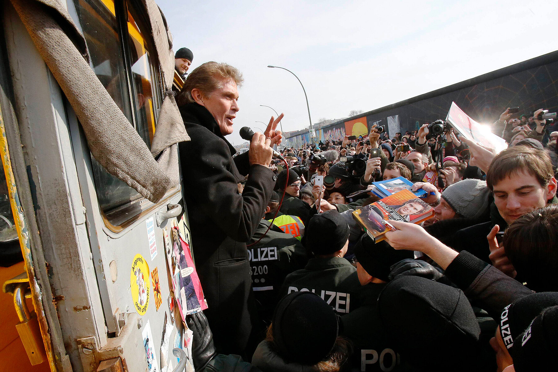 David Hasselhoff is standing in the doorway of a bus with a microphone in his hand. He is surrounded by demonstrators and police, with the East Side Gallery behind him on the right.