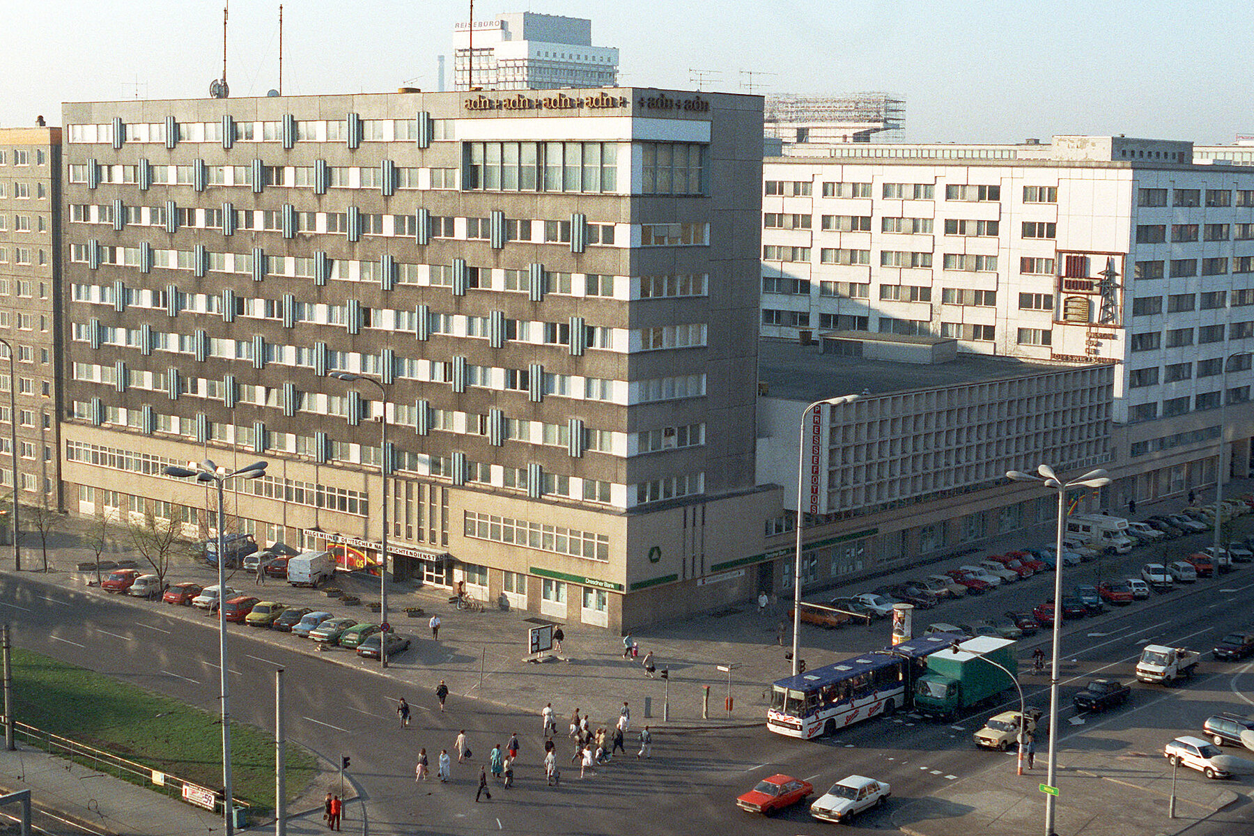 Grey prefabricated building with ADN lettering at the top right of the façade. In front of it an intersection where cars stop at traffic lights and people cross the street.