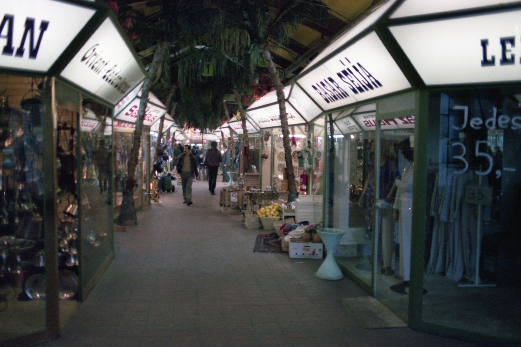 Various shops with household goods, clothing and food inside the Turkish Bazaar on the left and right side. People walk through the central aisle.