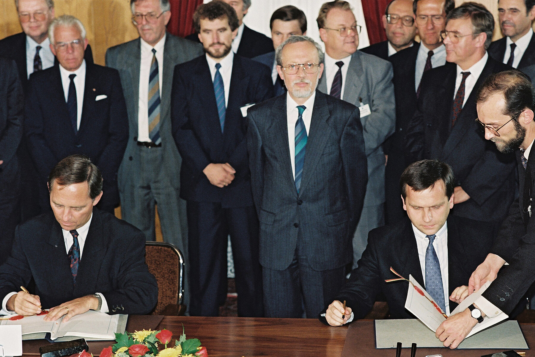 Several men in suits stand in a row. In the foreground, two men sit at a table, each signing a document.  