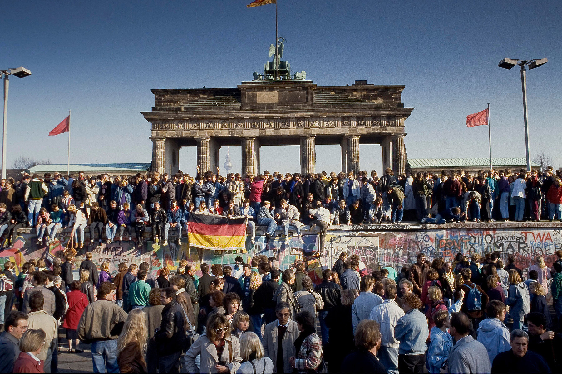People in front of the Brandenburg Gate and on top of the Berlin Wall during the daytime. In the centre of the picture, people hold up the German flag. 
