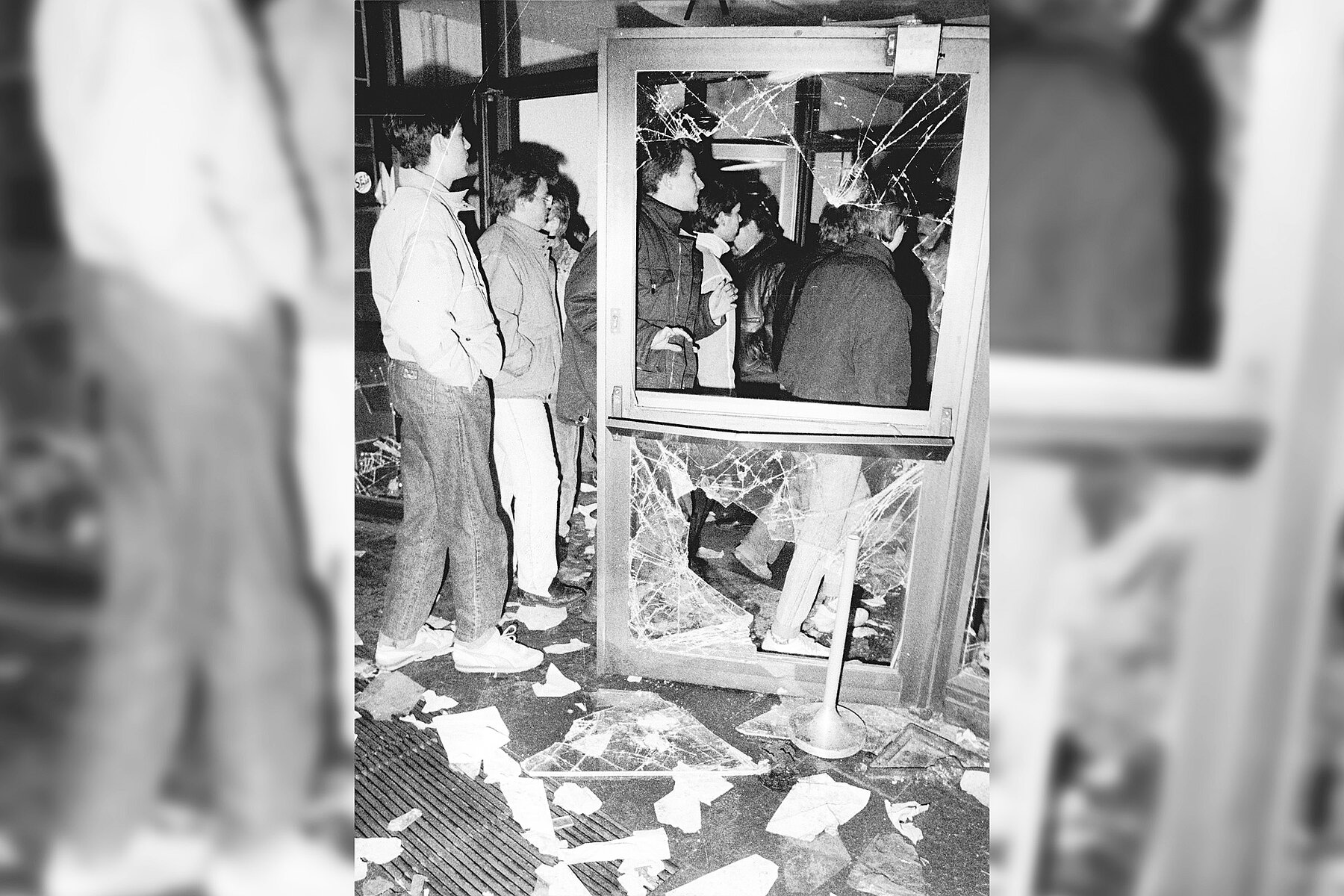 Six people stand in a building next to and behind an open door with completely smashed glass windows. There is paper and broken glass on the floor. 