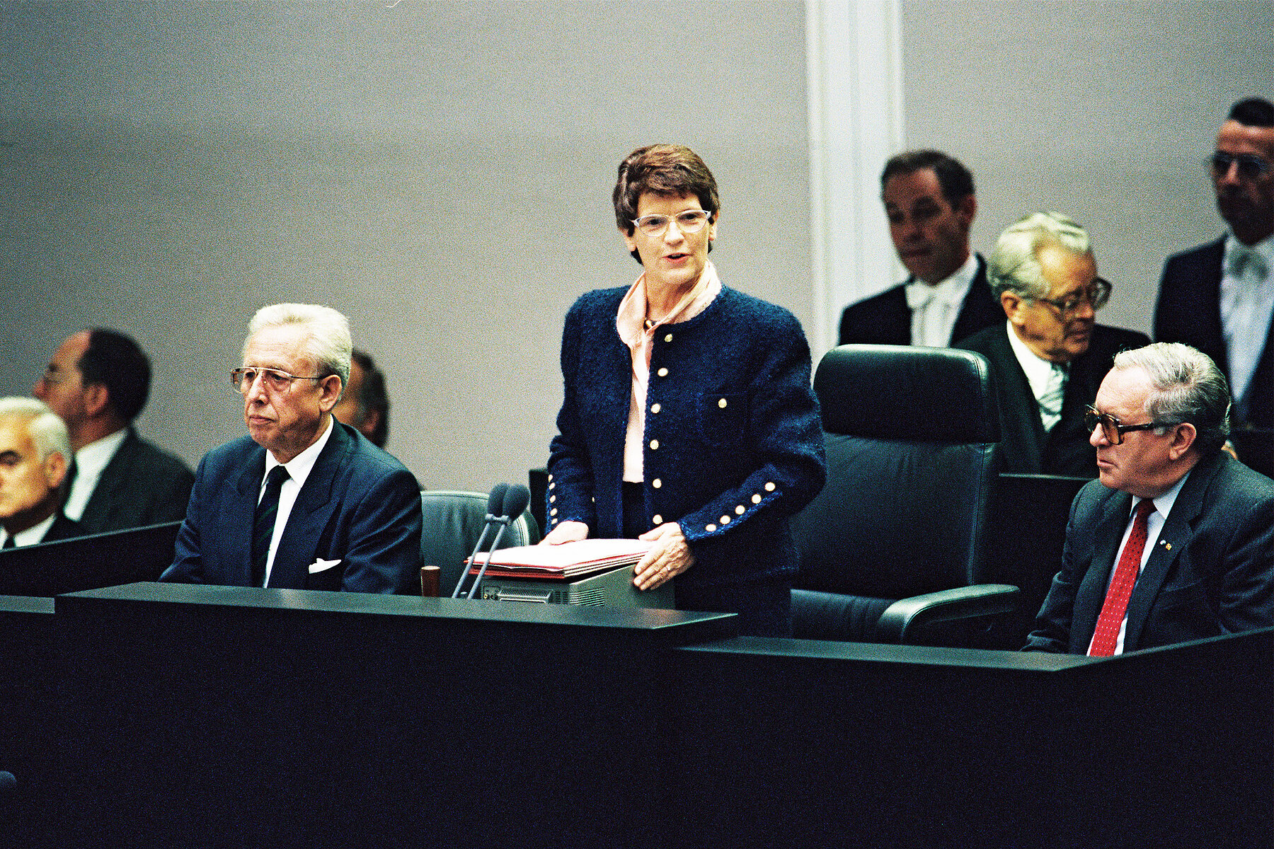 Rita Süssmuth steht an einem Podium und spricht umgeben von sitzenden männlichen Politikern. 