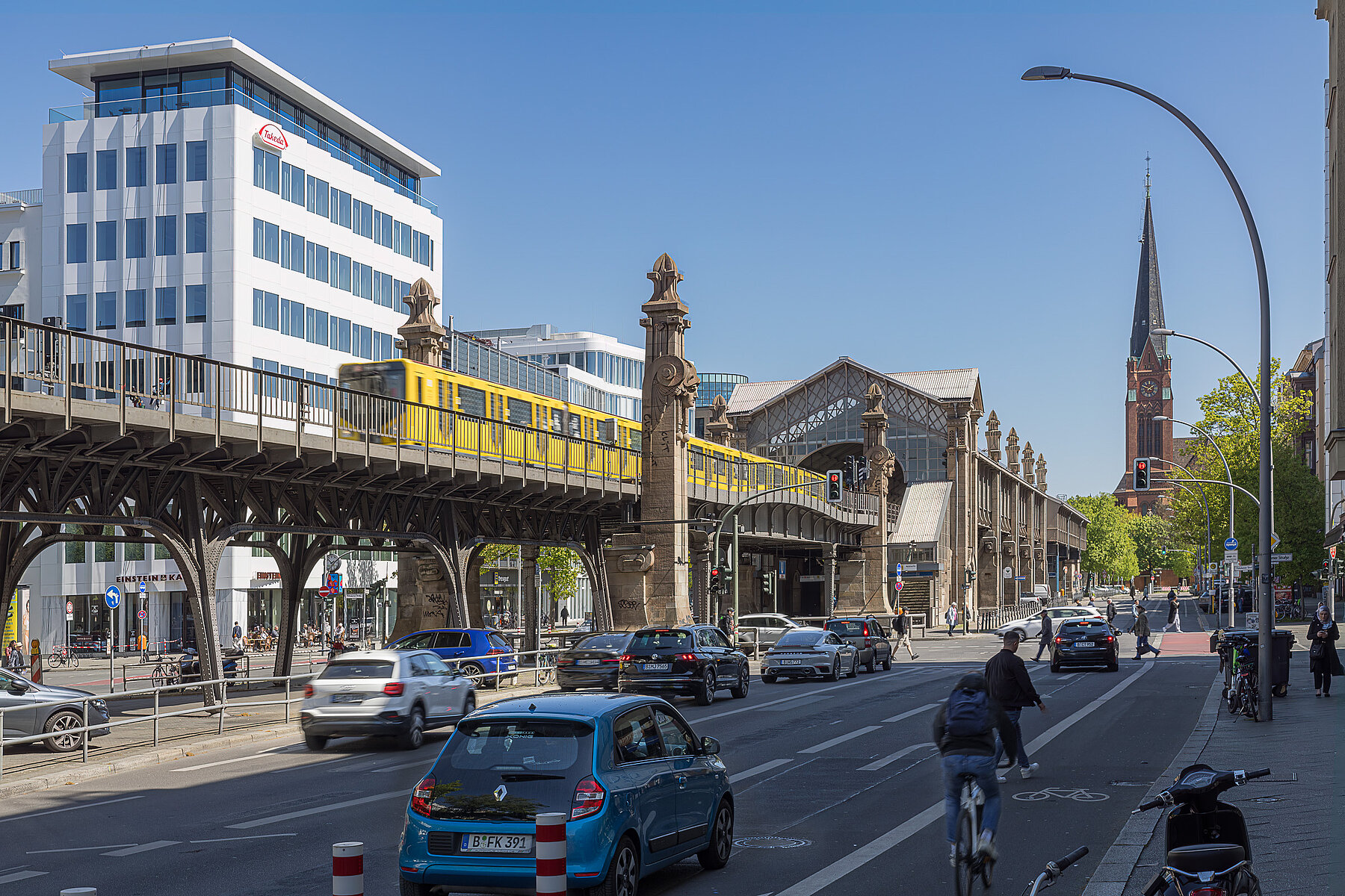 Eine gelbe U-Bahn fährt aus dem überirdisch gelegenen Bahnhof Bülowstraße, rechts daneben befindet sich eine Straße mit Autos und Radfahrern.