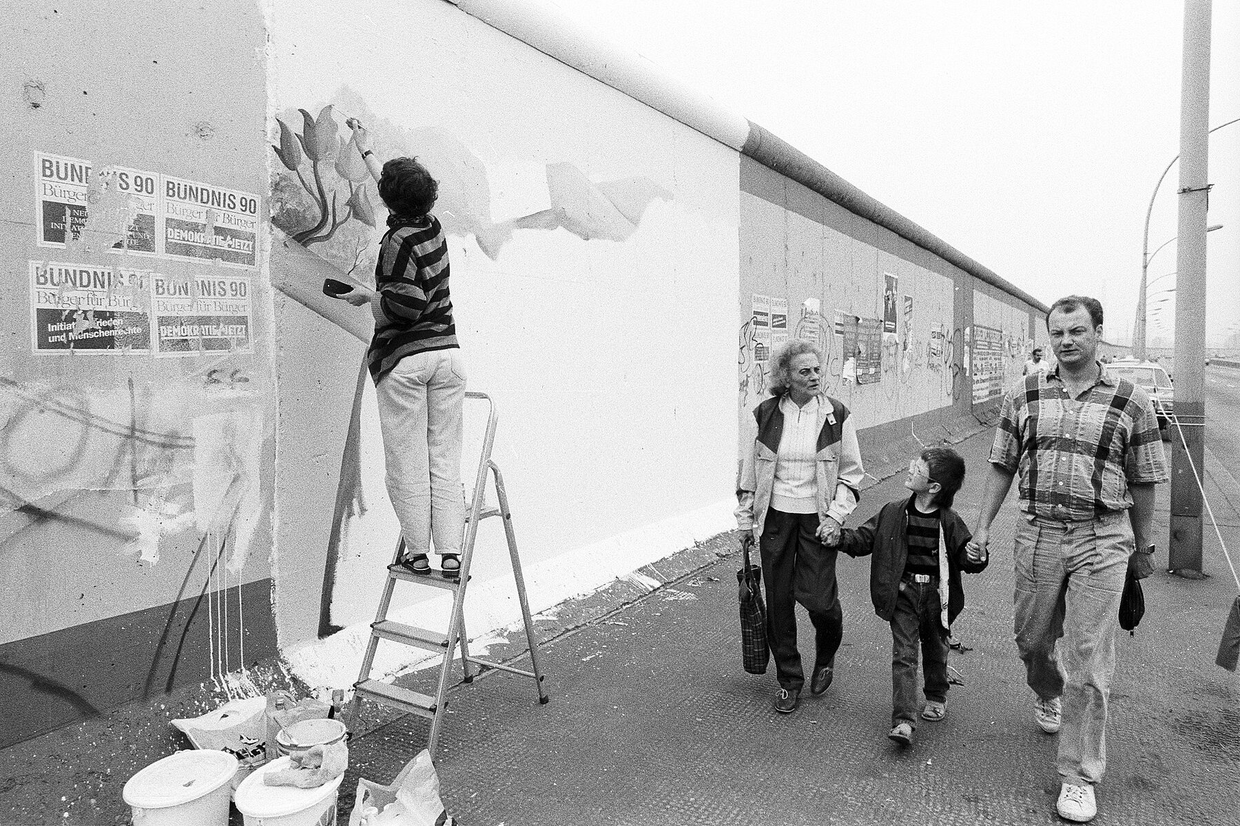 Links eine Person auf einer Leiter, die ein großes Bild auf die Mauer malt. rechts geht eine Familie mit einem Kind vorbei.