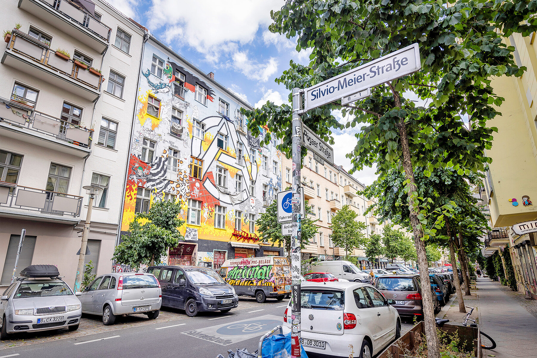 A street sign with the inscription Silvio-Meier-Straße. In the background is the colourfully painted house of a housing project at Rigaer Straße.
