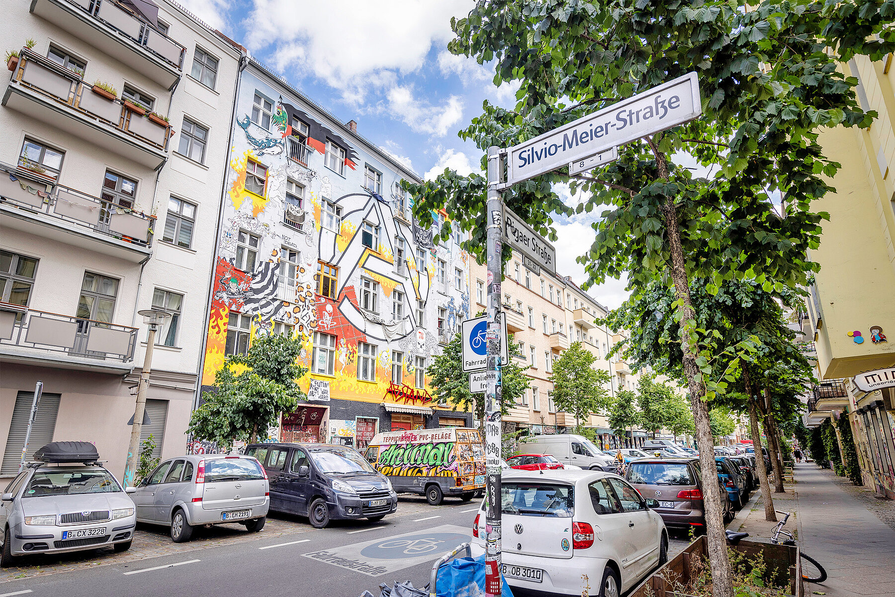 Straßenschild mit Aufschrift Silvio-Meier-Straße. Im Hintergrund das buntbemalte Haus eines Wohnprojekts an der Rigaer Straße.