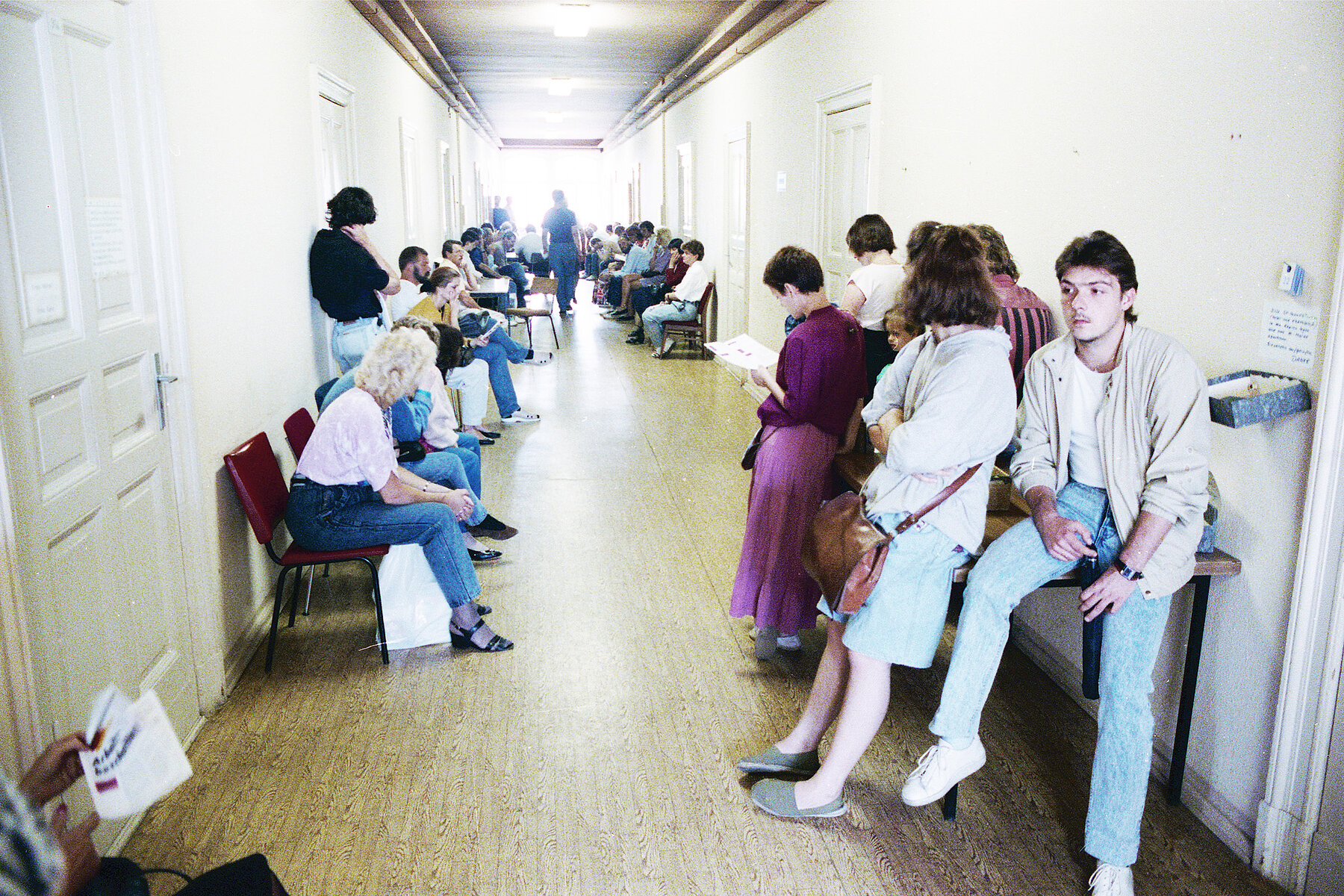 People wait in a beige office corridor, sitting and standing.
