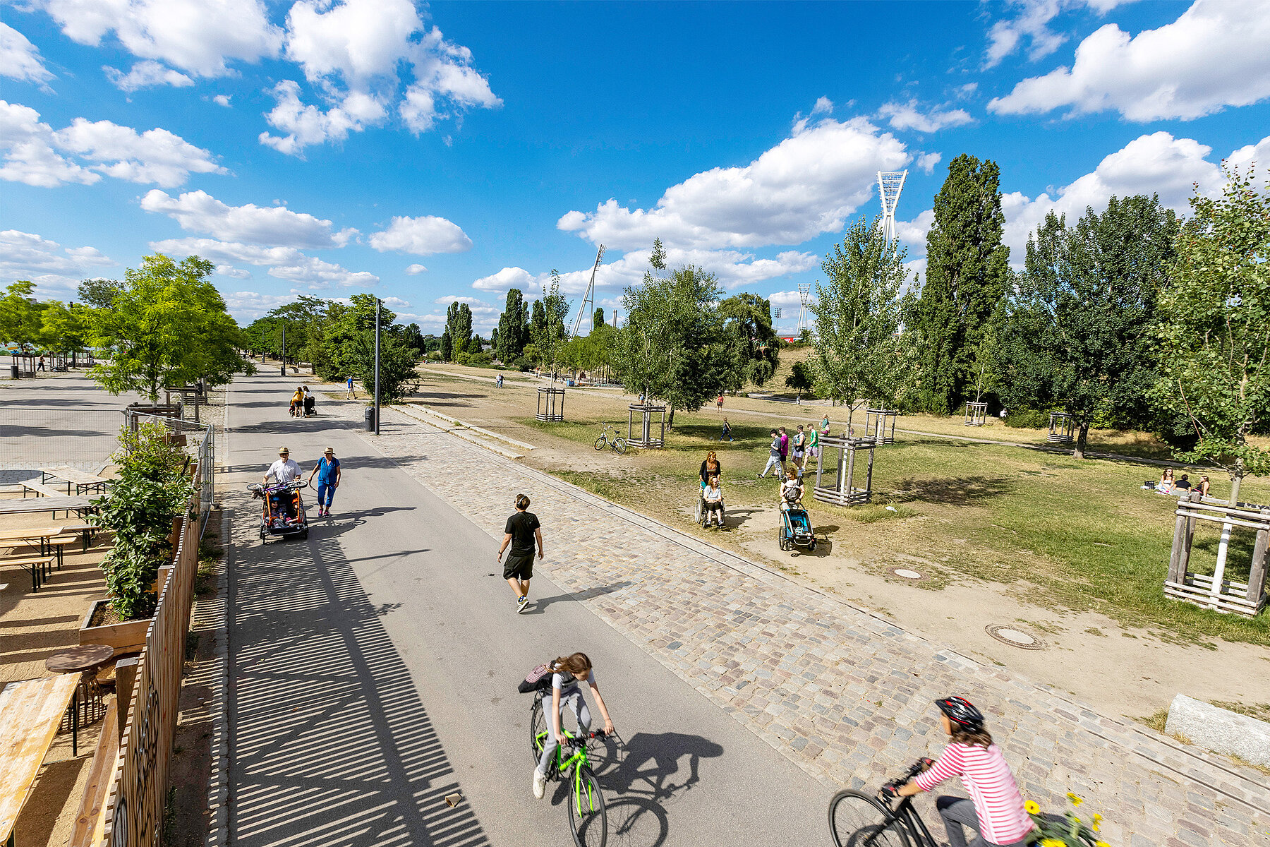 Menschen laufen auf einem asphaltierten Weg durch einen Park. Rechts ist eine Wiese mit Bäumen. Im Hintergrund befindet sich die Beleuchtung eines Stadions. Auf der linken Seite stehen Bierbänke.