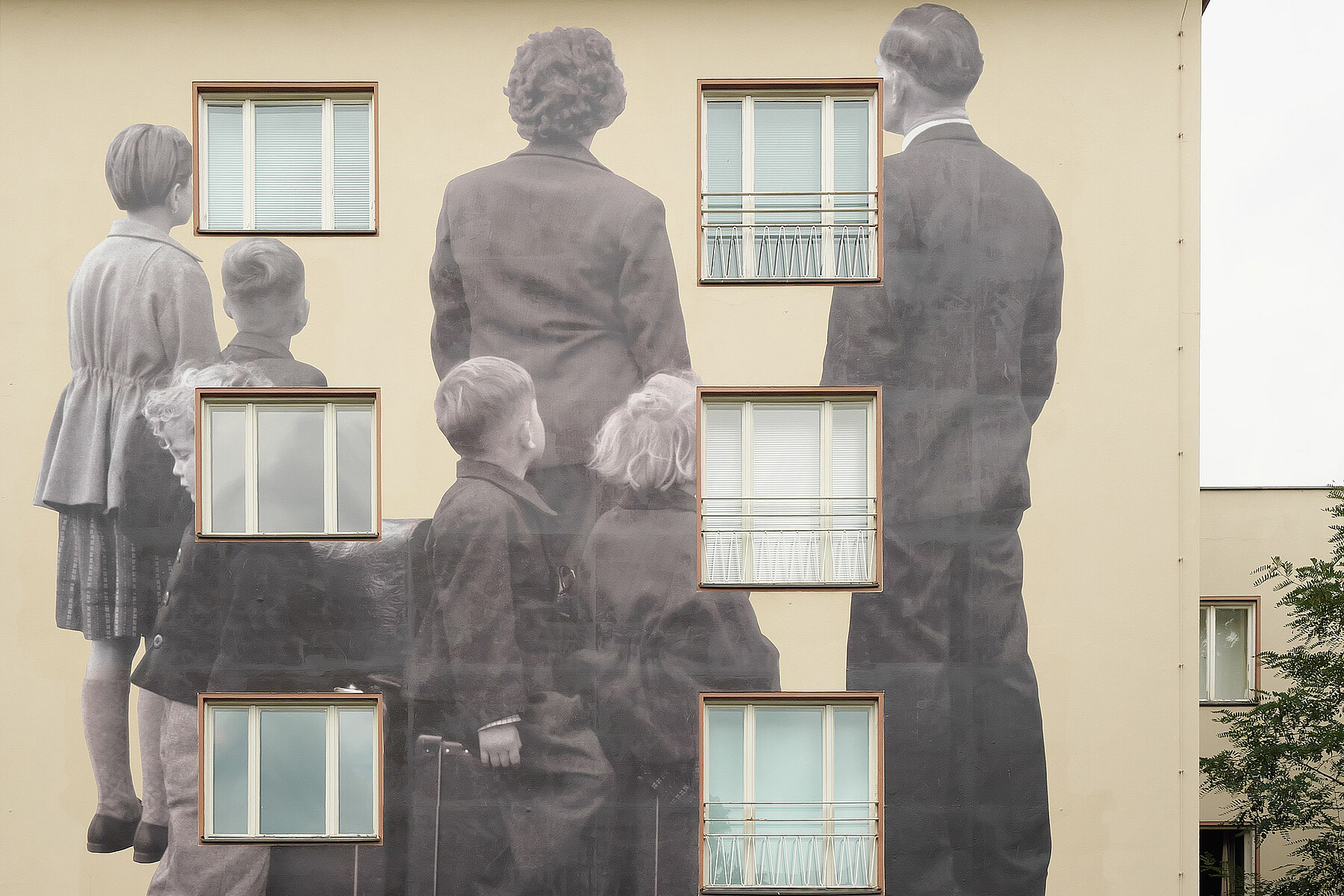 An einer Fassade befindet sich ein Wandbild, das über drei Stockwerke reicht. Dargestellt ist eine Familie in schwarzweiß bestehend aus Frau, Mann und vier Kindern. 