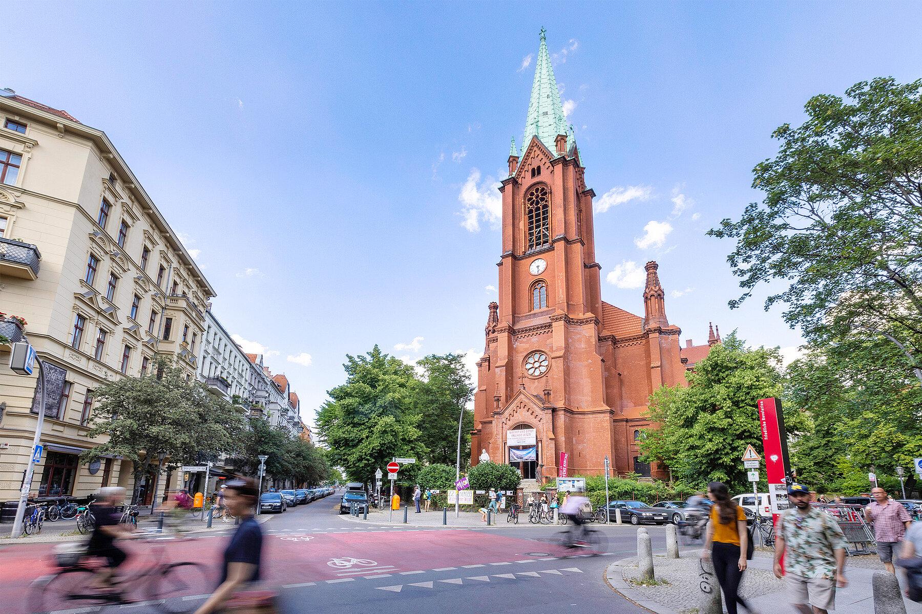 The Gethsemanekirche made of red bricks is located at a street crossing. People pass by on foot or on bicycles.