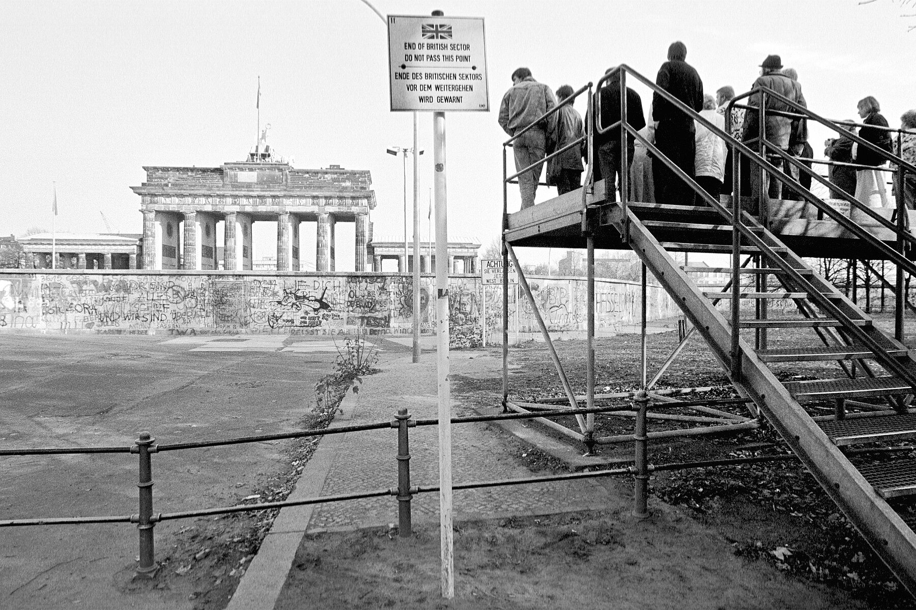 Menschen stehen auf einer Aussichtsplattform in Westberlin und blicken auf das Brandenburger Tor links hinter der Mauer. Im Vordergrund steht auf einem Schild auf Englisch und Deutsch: Ende des britischen Sektors. Vor dem Weitergehen wird gewarnt! 