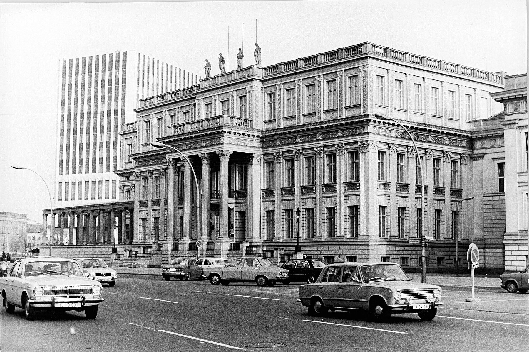 View of the Crown Prince's Palace. In front of it is a street with cars. In the background is the white high-rise building of the Ministry of Foreign Affairs of the GDR.