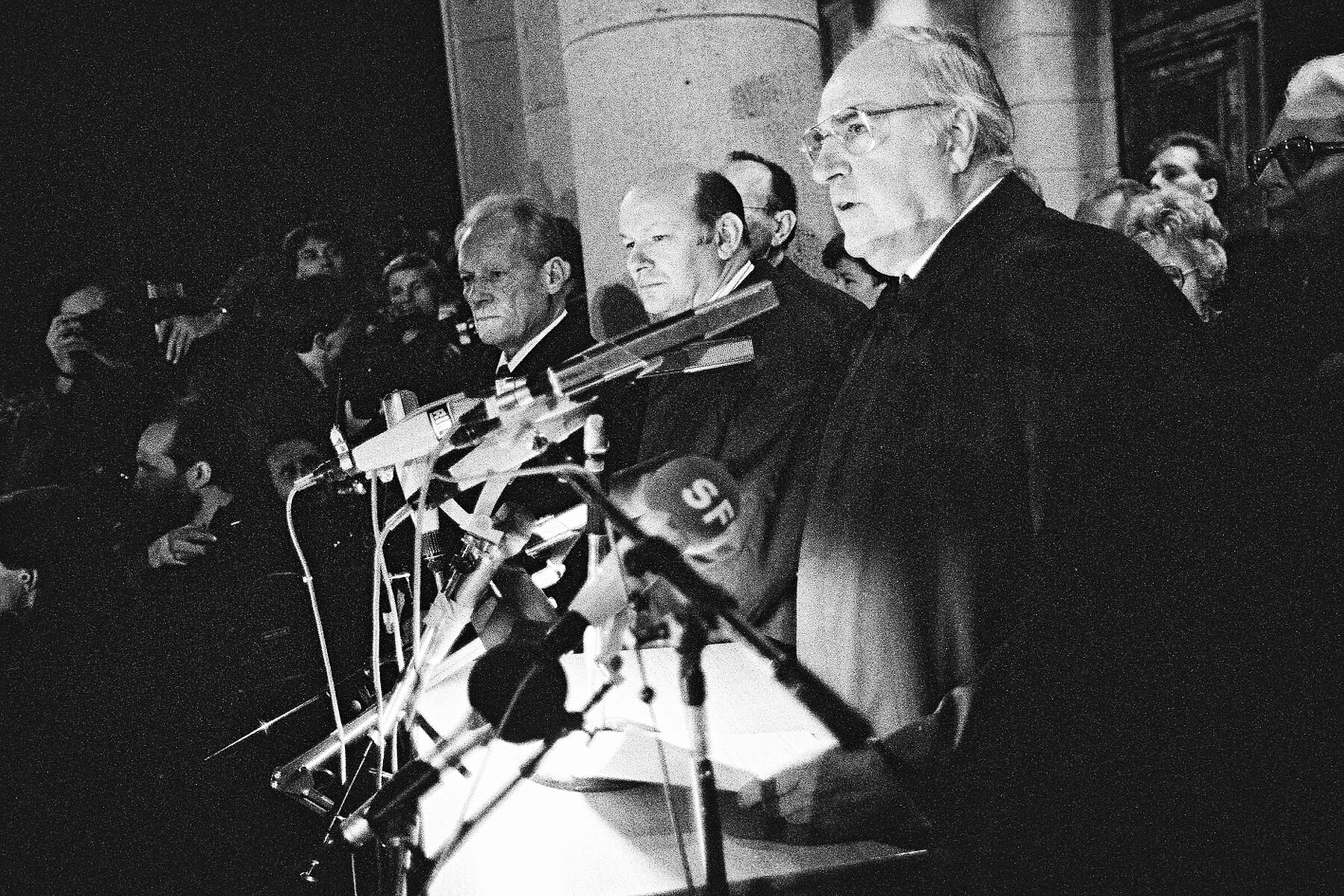 Mehrere Politiker stehen an einem Rednerpult vor Mikrofonen. Im Hintergrund fotografiert die Presse, im Vordergrund spricht Helmut Kohl. 