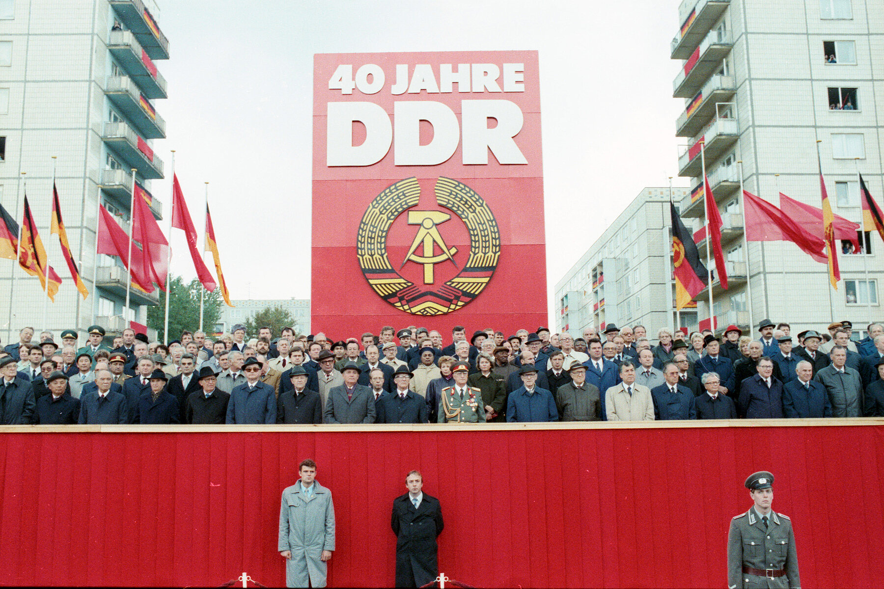 A group of men and a few women in suits and military uniforms stand in four rows on a grandstand. Behind the grandstand is a red sign that reads: 40 Years GDR. Below the inscription is the national emblem of the GDR.