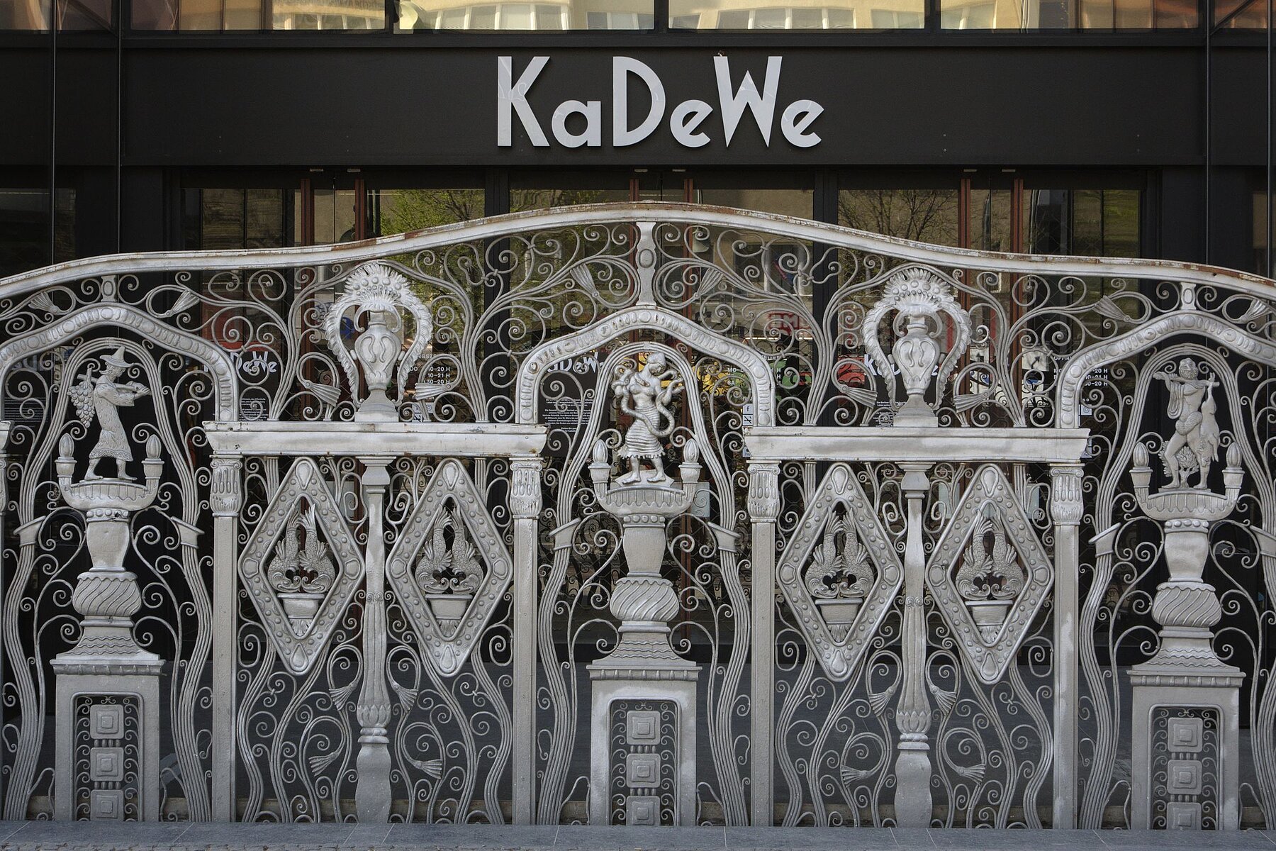 Cast-iron, silver-coloured gate with ornamental decorations. Above it hangs the KaDeWe lettering in white letters on a black background.