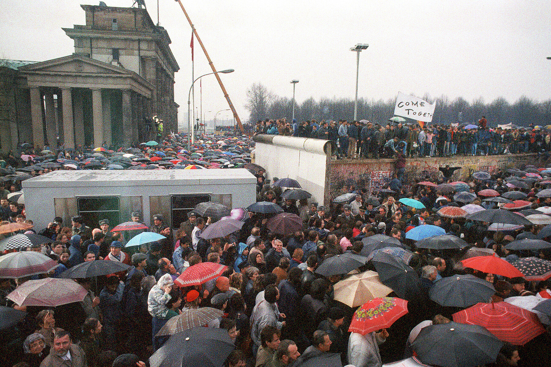 A container located to the side of the Brandenburg Gate is surrounded by many people with umbrellas. People on top of a Wall section hold up a sign that reads Come together.