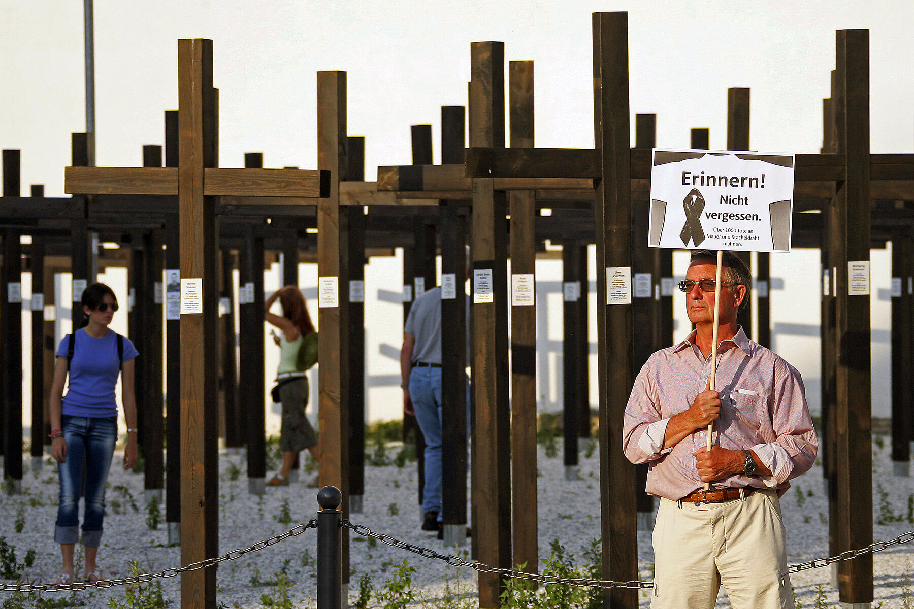 A man holds a sign that reads: Remember! Do not forget. More than 3,000 dead at the Wall and barbed wire remind us. In the background are several rows of large, upright wooden crosses. 