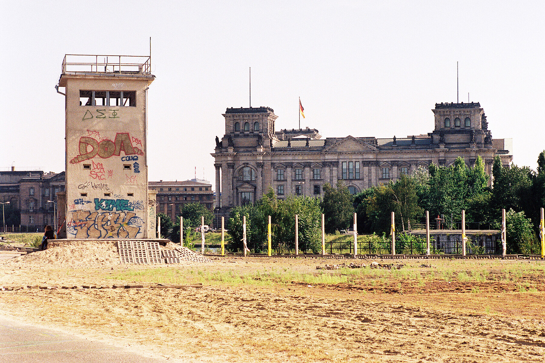 Links steht ein verlassener Wachtturm der Berliner Mauer, dahinter die Spree und das Reichstagsgebäude. 
