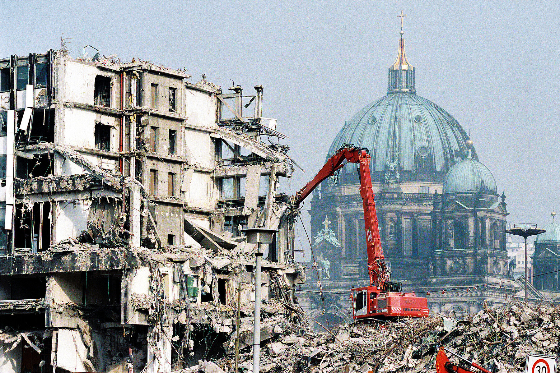 On the left, the high-rise building of the Ministry of Foreign Affairs of the GDR is being demolished by a red excavator. Behind it on the right is the Berlin Cathedral.  