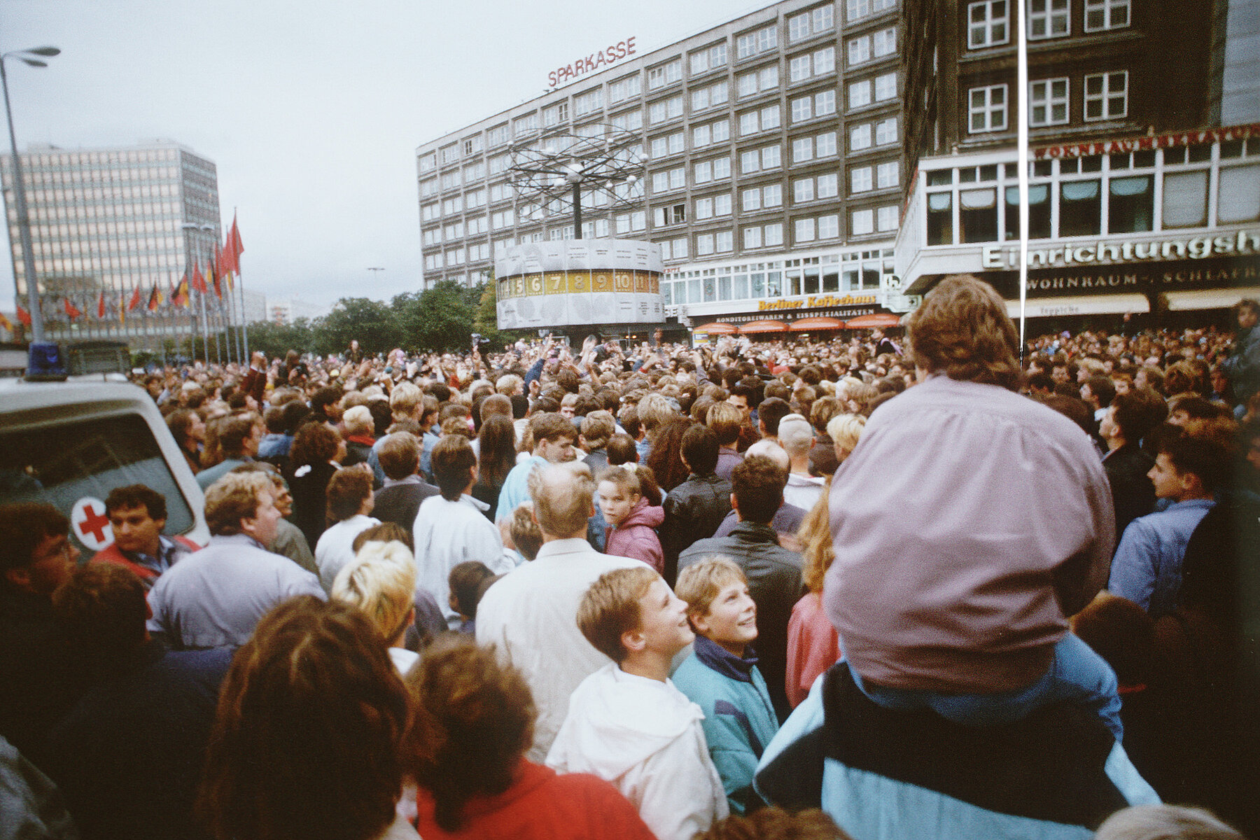 A crowd of people at Alexanderplatz. In the background on the left is the House of the Teacher, in the middle the World Clock.