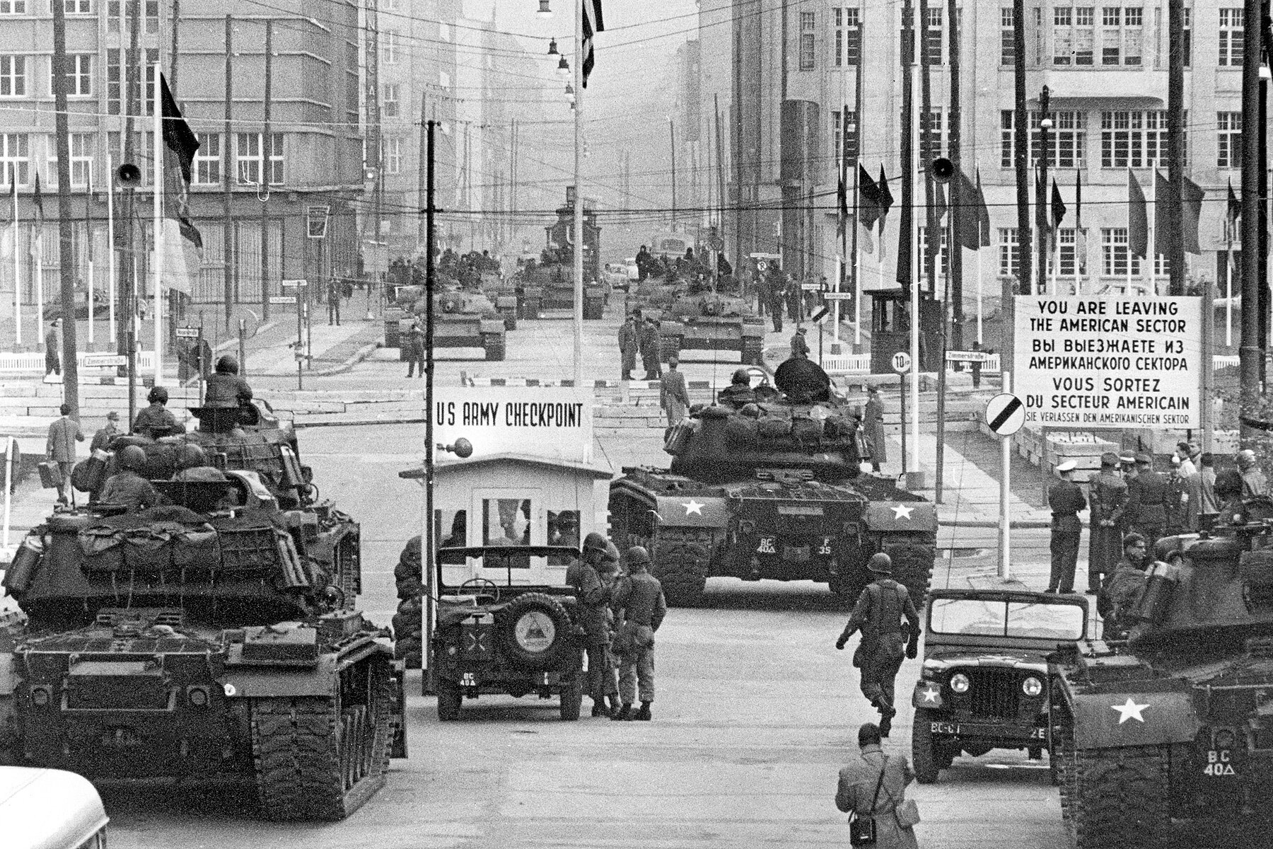 Am Checkpoint Charlie stehen sich Panzer mit Soldaten gegenüber. Mehrere Flaggen sind links und rechts gehisst. Rechts im Hintergrund steht auf einem Schild: Sie verlassen den Amerikanischen Sektor, auf Englisch, Russisch, Französisch und Deutsch.