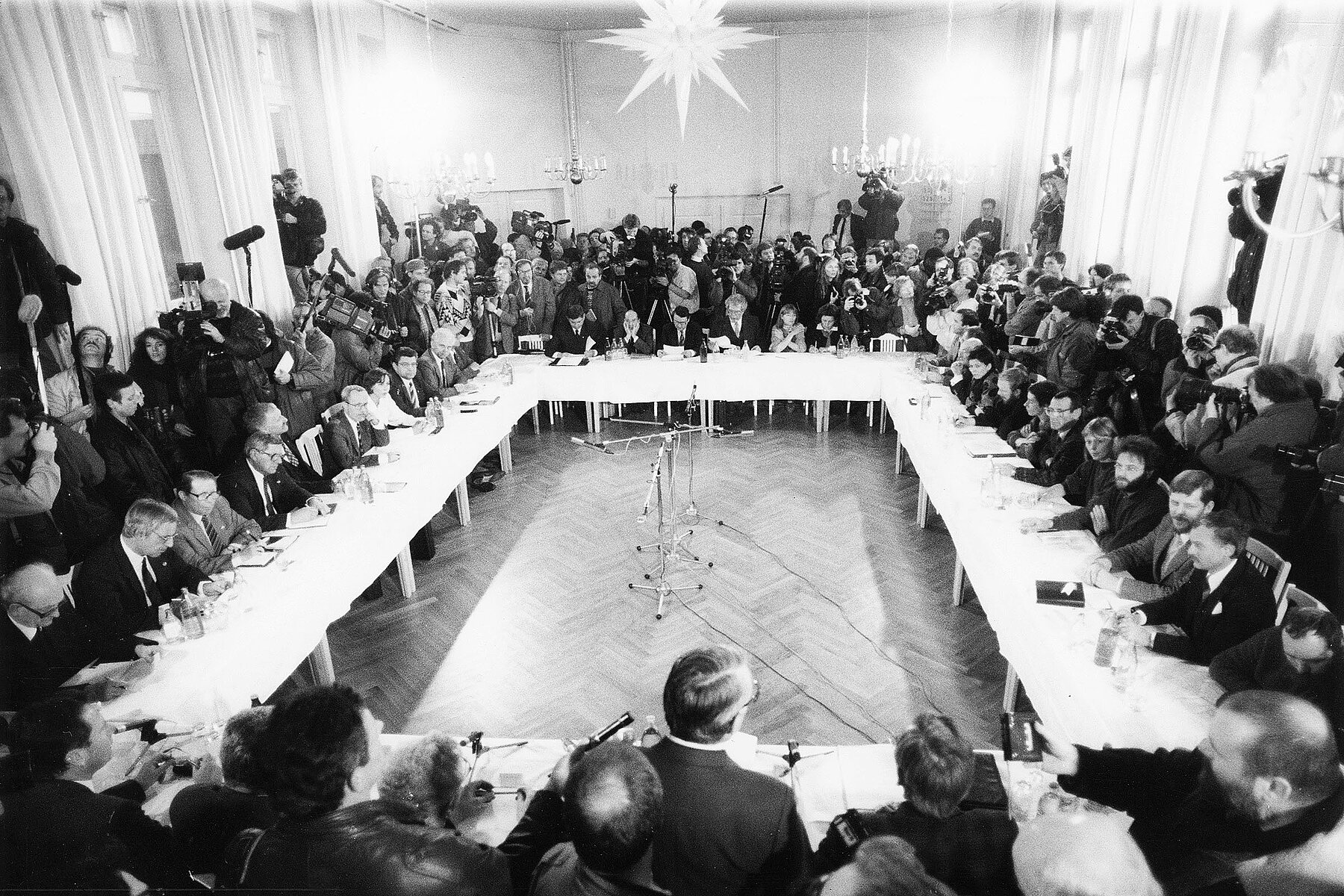 Participants seated at conference tables set up in a square at the first meeting of the Round Table in a house of the church, with spectators and media people standing around the table.