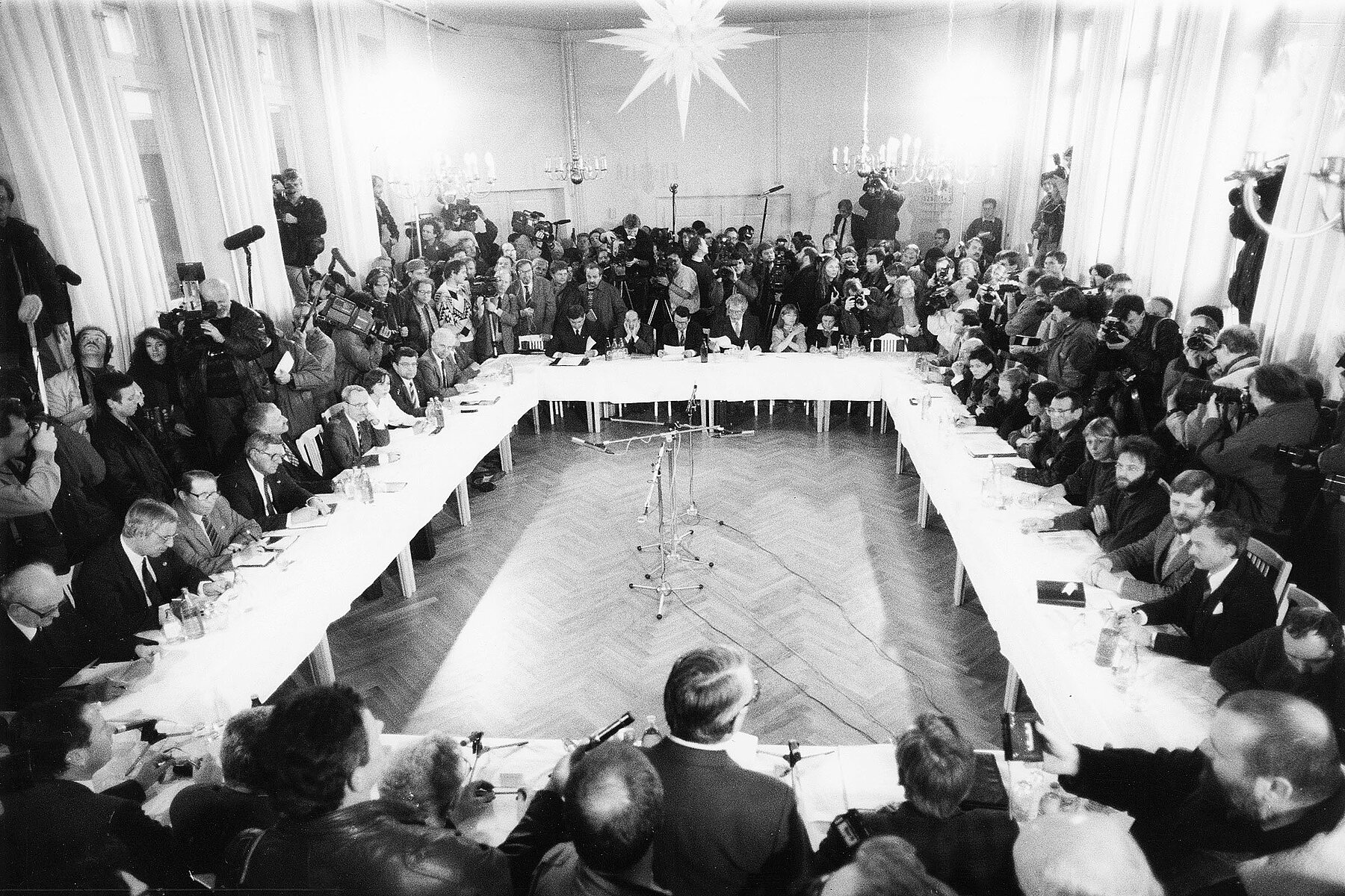 Teilnehmende sitzen bei der ersten Sitzung des Runden Tisches in einem Haus der Kirche, an im Viereck aufgestellten Konferenztischen, um den Tisch herum stehen Zuschauende und Medienleute.